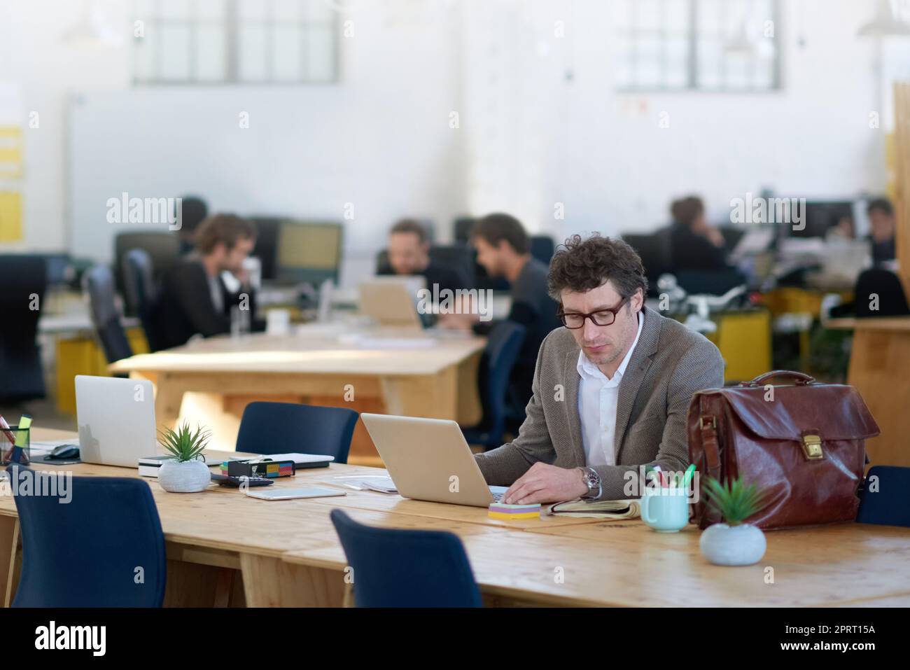 Wenn Technologie eine Sprache wäre, dann würde er fließend sein. Ein reifer Mann, der in einem großen Büro mit Kollegen im Hintergrund an einem Laptop arbeitet. Stockfoto