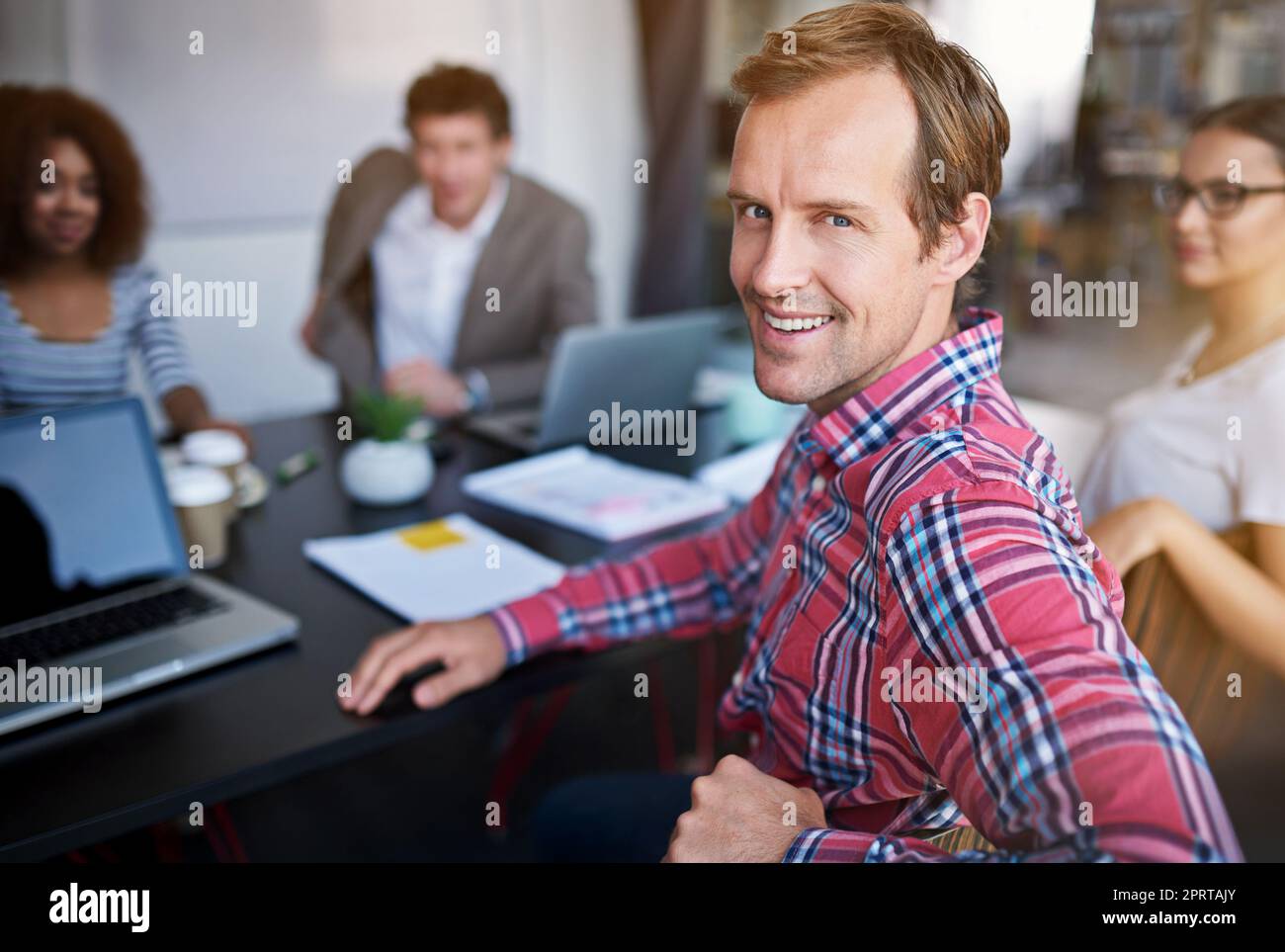 In diesem Büro werden Ideen zum Leben erweckt. Porträt eines Büromitarbeiters, der an einem Sitzungstisch sitzt, mit Kollegen im Hintergrund. Stockfoto