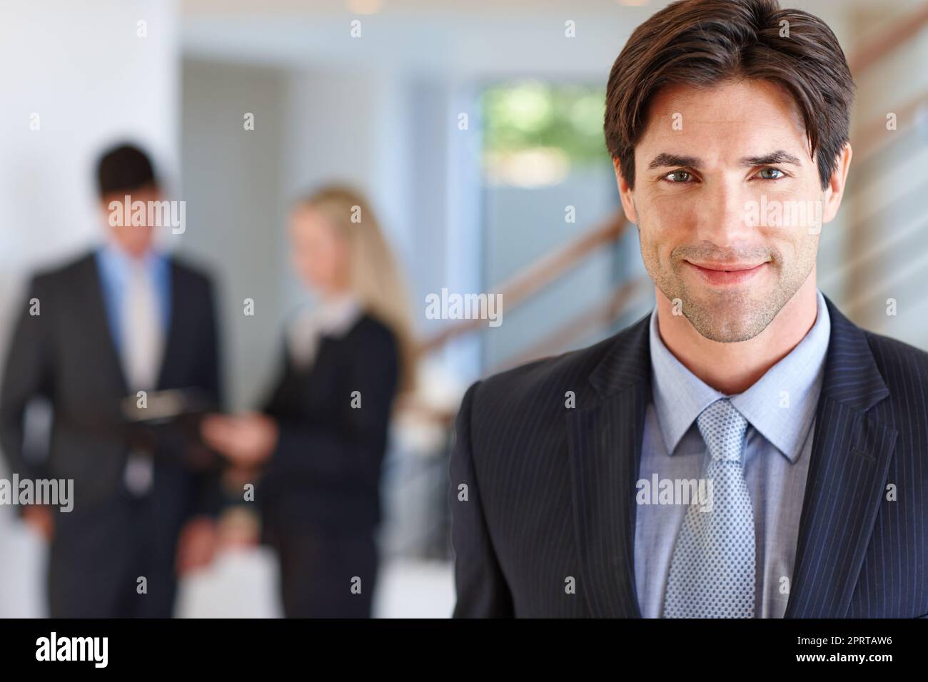 Das Geschäft kommt ihm natürlich zu. Porträt eines Geschäftsmannes, der in einem Gebäude mit zwei Kollegen im Hintergrund steht. Stockfoto