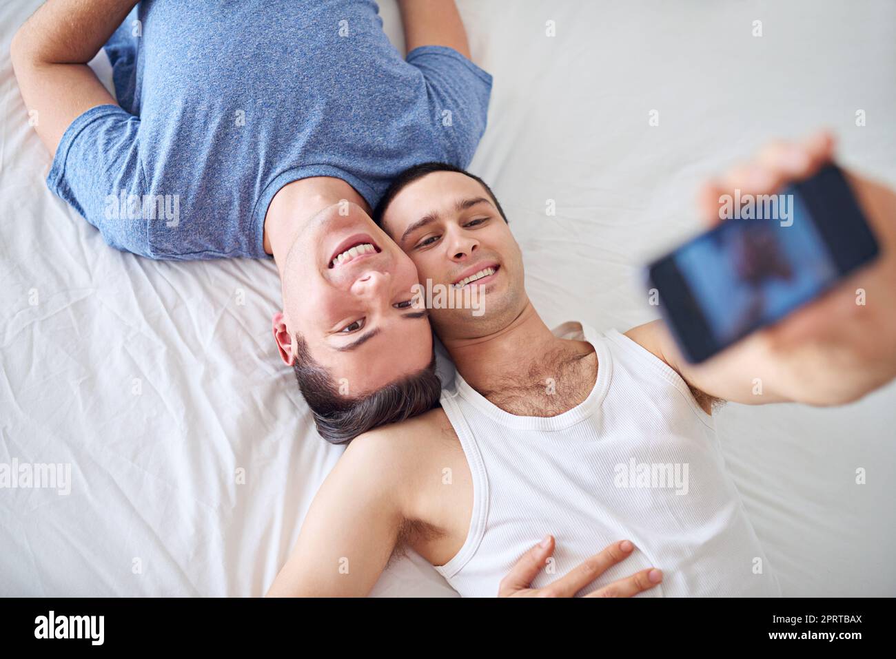 Machen Sie es zu einem Keeper mit einem großen Lächeln. Ein junges schwules Paar, das ein Selfie macht, während es sich im Bett entspannt. Stockfoto