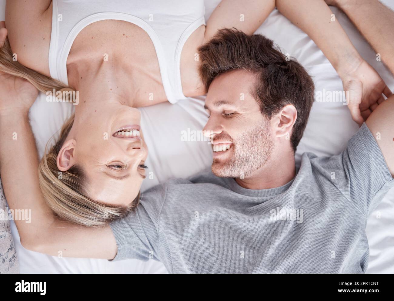 Weißes glückliches Paar oben, Lächeln und Bett zusammen entspannen in Glück für Beziehung, Bindung und Pflege zu Hause. Mann und Frau lächeln in Liebe, liegen und entspannen für Schlafenszeit und Pause in Komfort Stockfoto