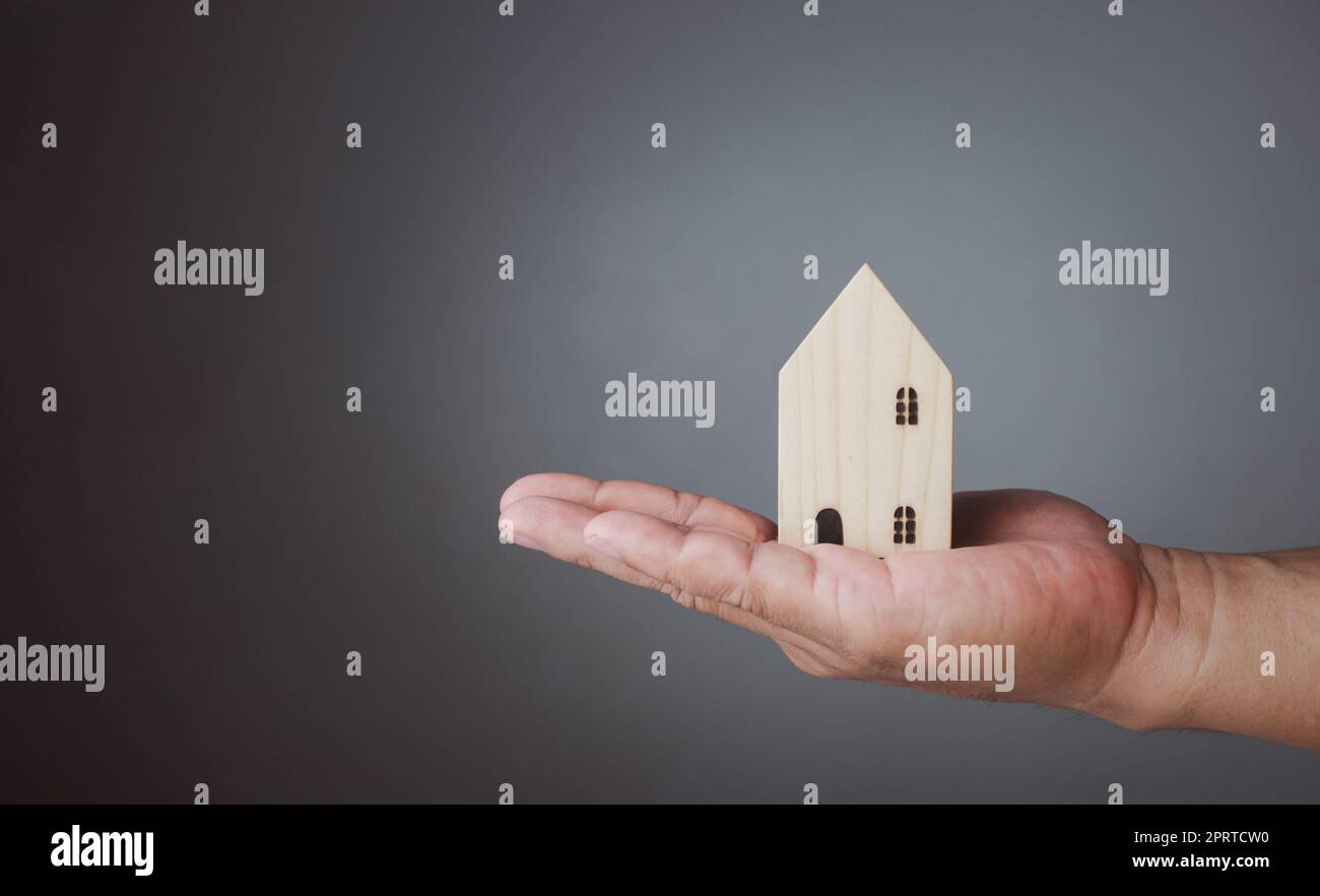 Konzept des Verkaufs eines Hauses. Eine Hand hält ein Modellhaus auf grauem Hintergrund. Immobilienmakler bieten Haus, Sachversicherung und Sicherheit, erschwingliche Wohnkonzepte, Hausversicherungsvermittler, Verkäufer. Stockfoto