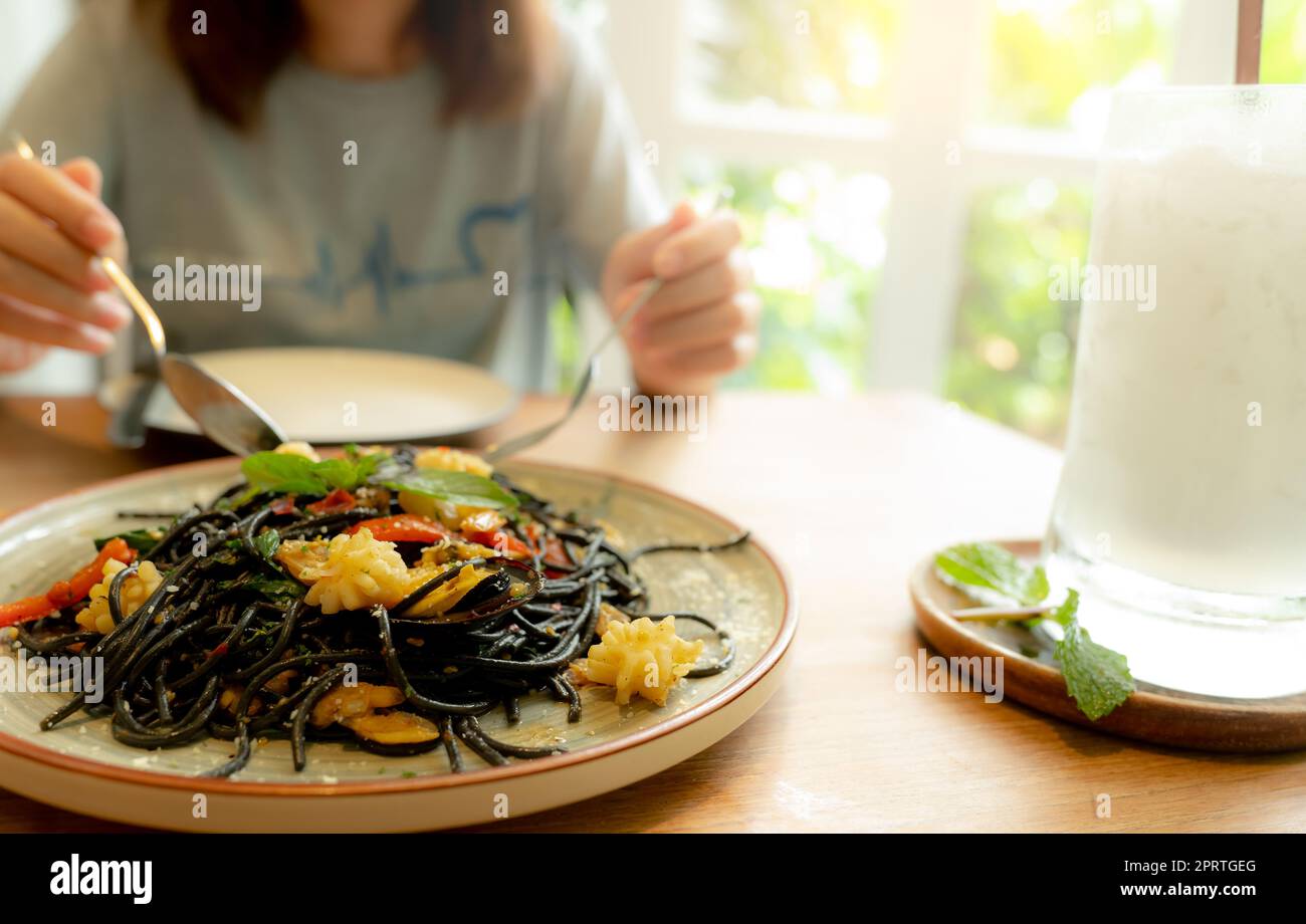 Schwarze Tintenfisch-Tintenfisch-Spaghetti mit Garnelen auf dem Teller. Schwarze Pasta mit Tintenfischtinte auf einem Restauranttisch und verschwommene Frau, die mit Gabel und Löffel isst. Gesundes Essen. Eine Frau isst köstliche schwarze Spaghetti. Stockfoto