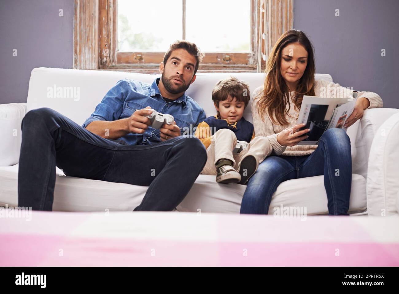 Zeit mit der Familie ist gut verbracht. Ein Sohn und sein Vater spielen zusammen ein Videospiel, während seine Mutter ein Magazin liest. Stockfoto