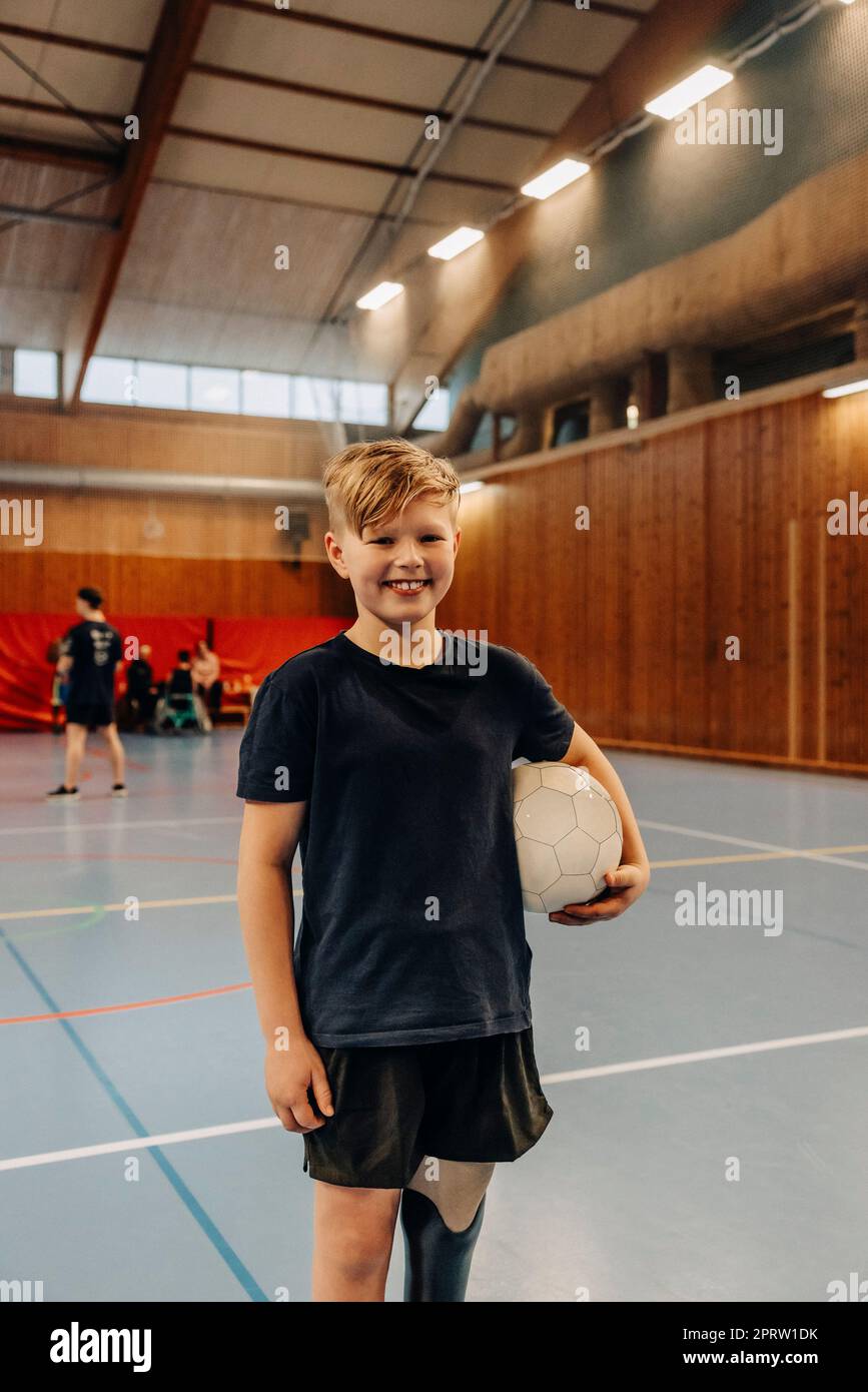Lächelnder Junge mit Behinderung hält Fußball, während er auf dem Sportplatz steht Stockfoto