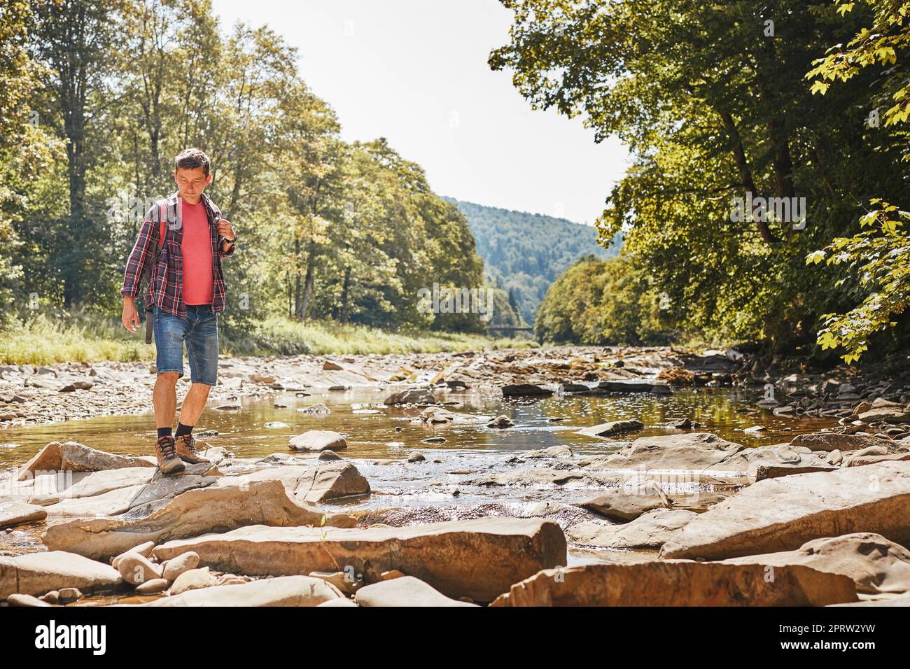 Trekking mit Rucksackkonzept. Rucksacktourist mit Wanderstiefeln über den Mountain River. Mann, der während des Sommerurlaubs in den Bergen wandert. Urlaub Stockfoto