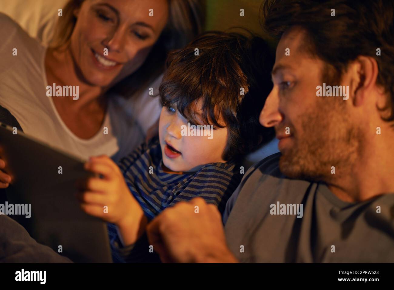 Familienfilmnacht im Bett. Eine junge Familie, die im Bett liegt und auf ein digitales Tablet schaut. Stockfoto