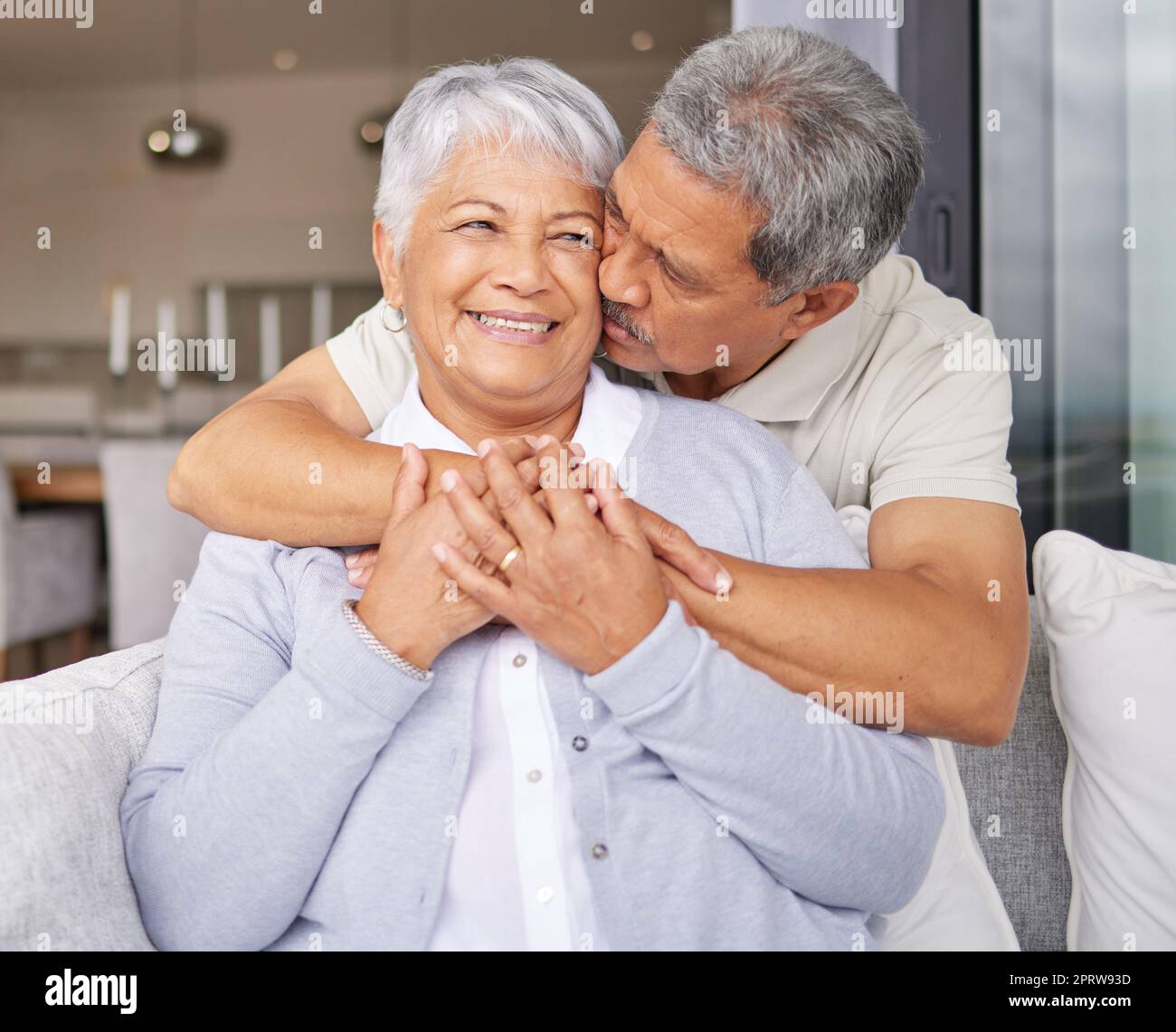 Glückliches Seniorenpaar küsst auf dem Sofa und lächelt für Liebe, Glück und Vertrauen in das Familienhaus oder das Wohnzimmer. Ältere Männer und Frauen aus Mexiko oder Leute im Ruhestand umarmen, küssen und romantisch auf der Couch Stockfoto