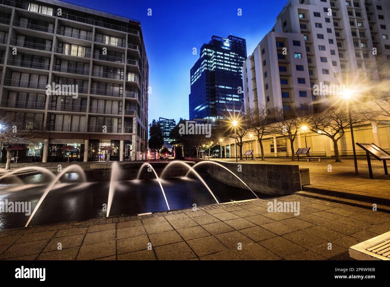Die Sonne geht über der Stadt unter. Ein Brunnen, der nachts auf einem leeren Stadtplatz fließt. Stockfoto
