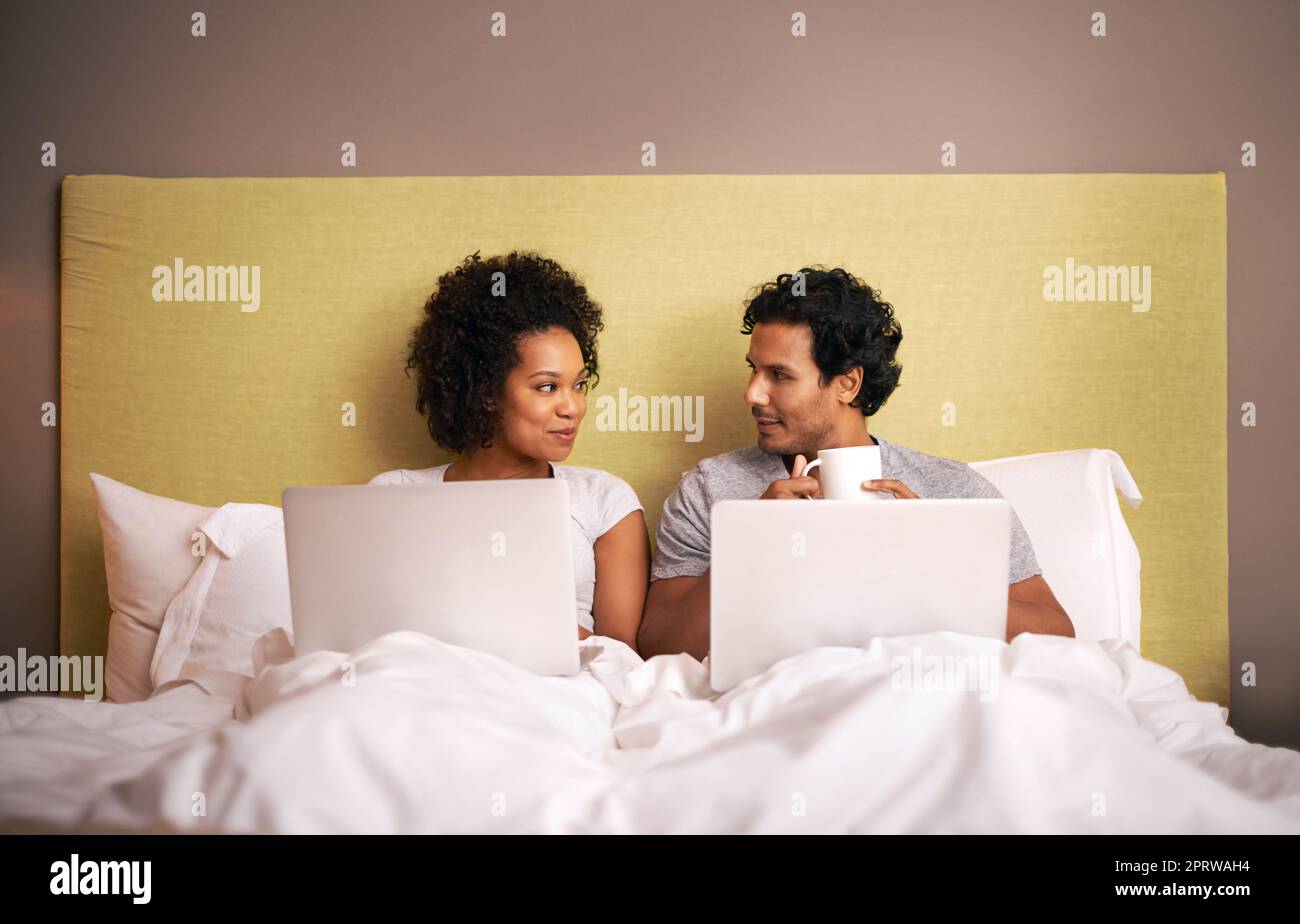 Arbeitet, während er sich unterhält. Ein junges Paar im Bett mit ihren Laptops. Stockfoto