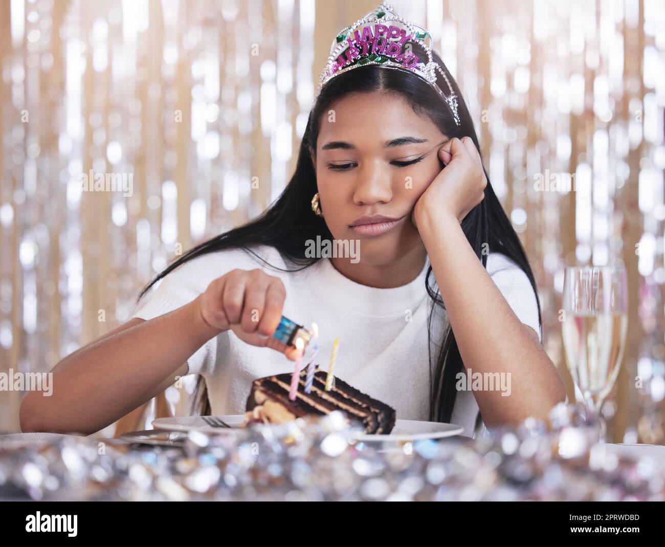 Traurige, depressive und wütende Frau bei der Geburtstagsfeier, während Kerzen auf Kuchen angezündet werden. Einsame, deprimierte und frustrierte Frau, die auf einer Party sitzt, versagt und einen Tisch mit Snack, Dekoration und Diadem serviert Stockfoto