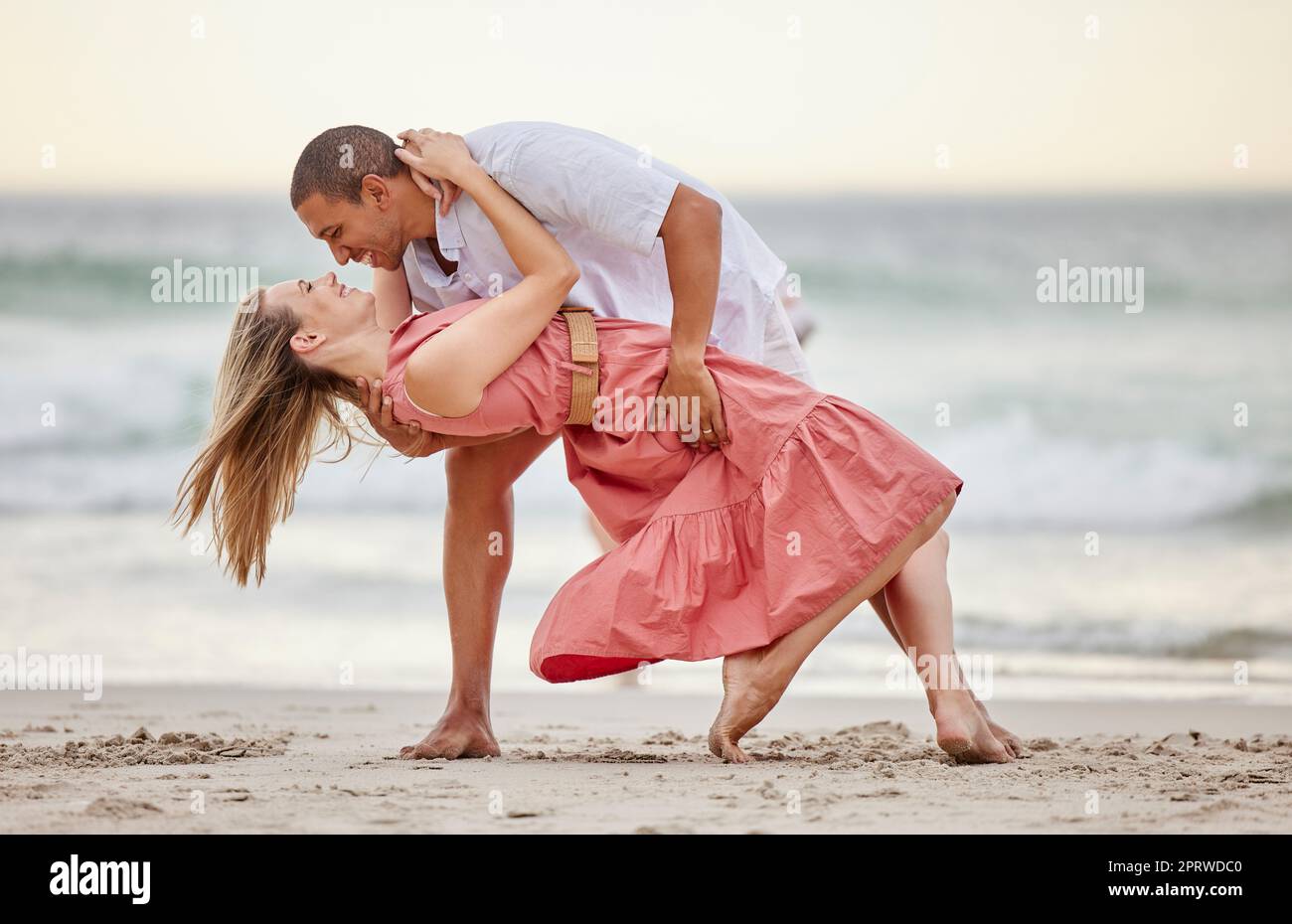 Liebe, Tanz und ein Paar, das im Sommer am Strand tanzt, feiern ihre Ehe, ihr Glück und ihre Flitterwochen. Lächeln, Sonnenuntergang und glückliche Frau, die eine gesunde Beziehung mit Partner auf See feiert Stockfoto