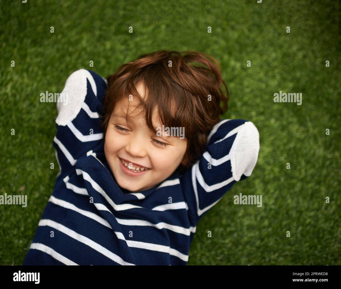 Tolles Kichern. Kleiner Junge, der auf dem Gras liegt, mit den Händen hinter dem Kopf. Stockfoto