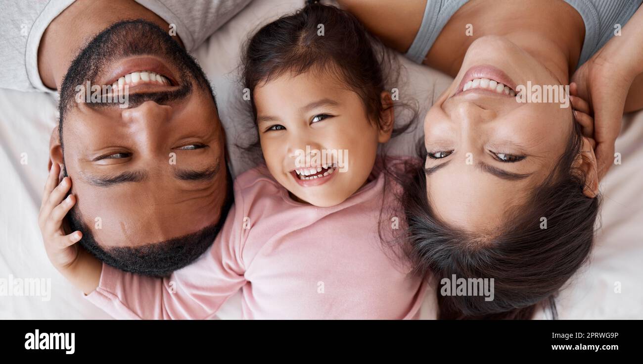 Glückliche Familie mit Kind im Bett in einem Gesichtsbild für zwischenrassische Liebe, Fürsorge und Glück zusammen. Mädchen aus Mexiko mit Mutter und Vater oder Eltern, die sich zu Hause entspannen und sich von oben verbinden Stockfoto