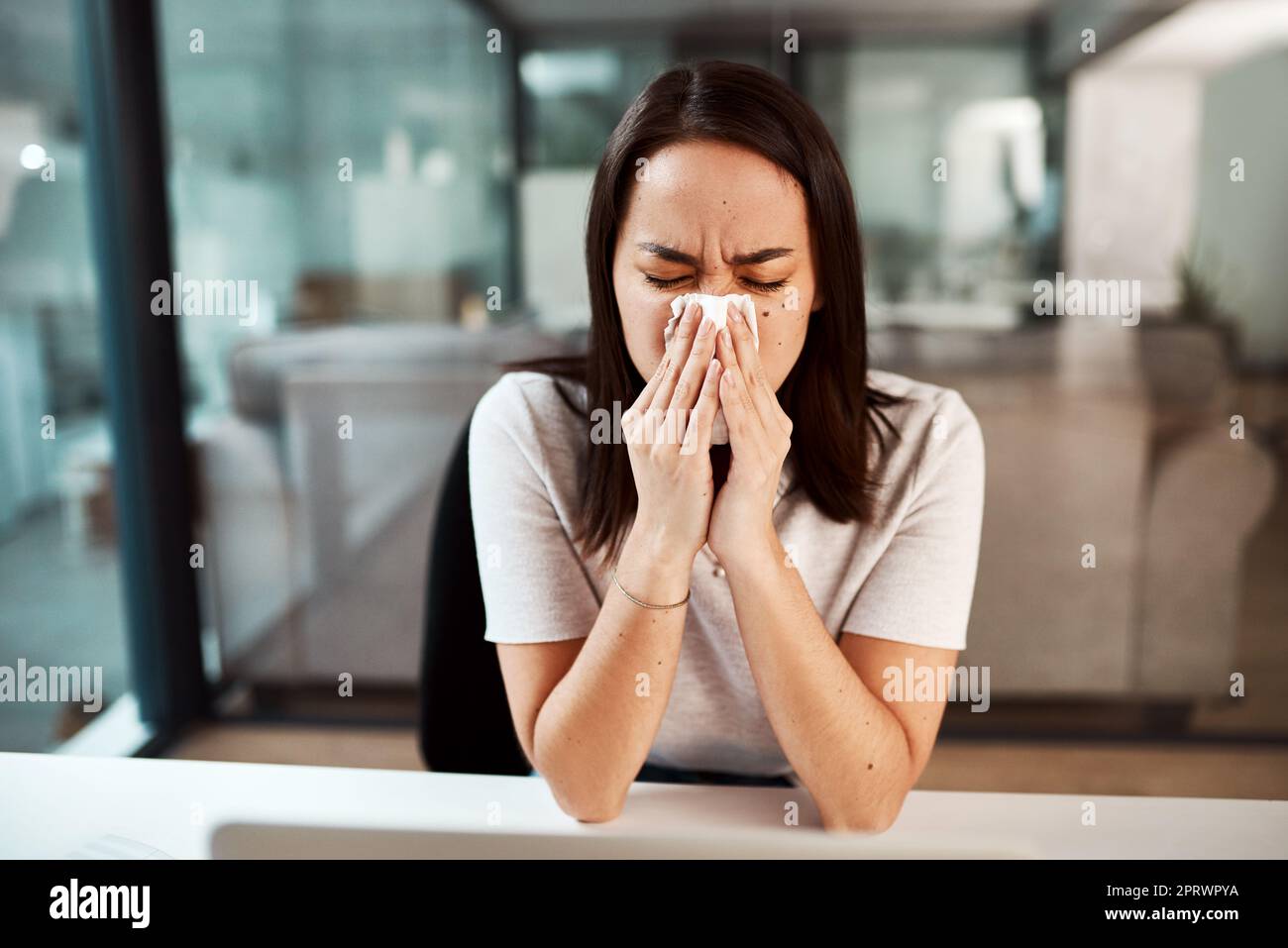 Der Umgang mit einer stickigen und laufenden Nase. Eine junge Geschäftsfrau bläst ihre Nase, während sie in einem Büro arbeitet. Stockfoto