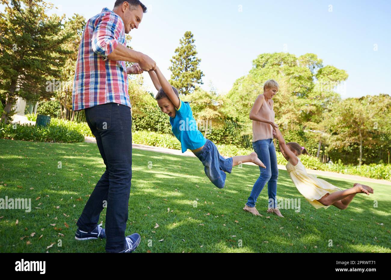 Wer braucht einen Spielplatz, wenn man mom und Dad um sich hat? Zwei Eltern schwingen ihre kleinen Kinder herum, während sie an einem wunderschönen sonnigen Tag im Park spielen. Stockfoto