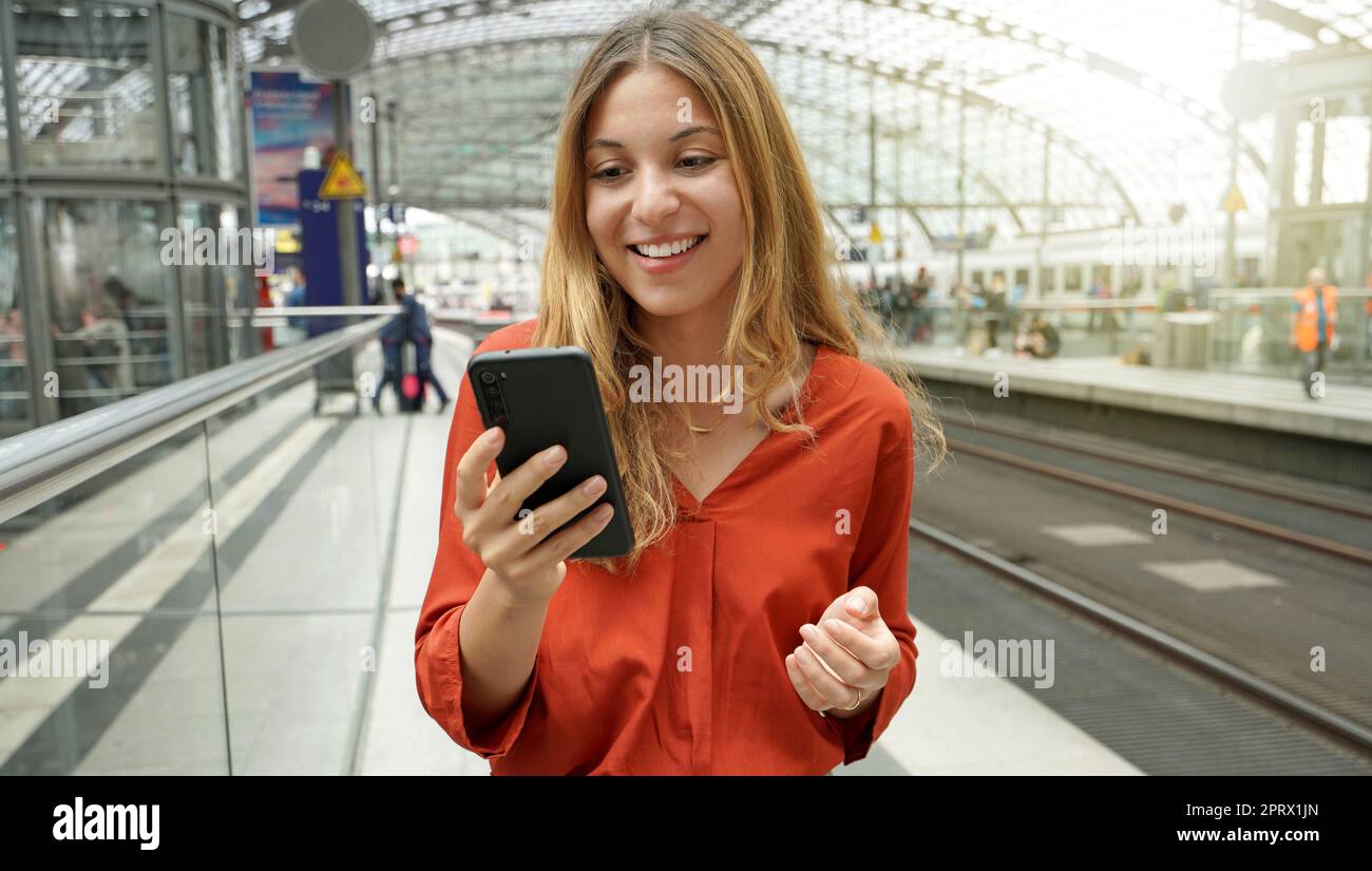 Junge lächelnde Frau, die ihr Handy auf der Sonnenstrahler-Plattform hält. Traveler Girl auf dem Bahnhof mit Handy. Digitales WiFi-Internet-Lifestyle. Stockfoto