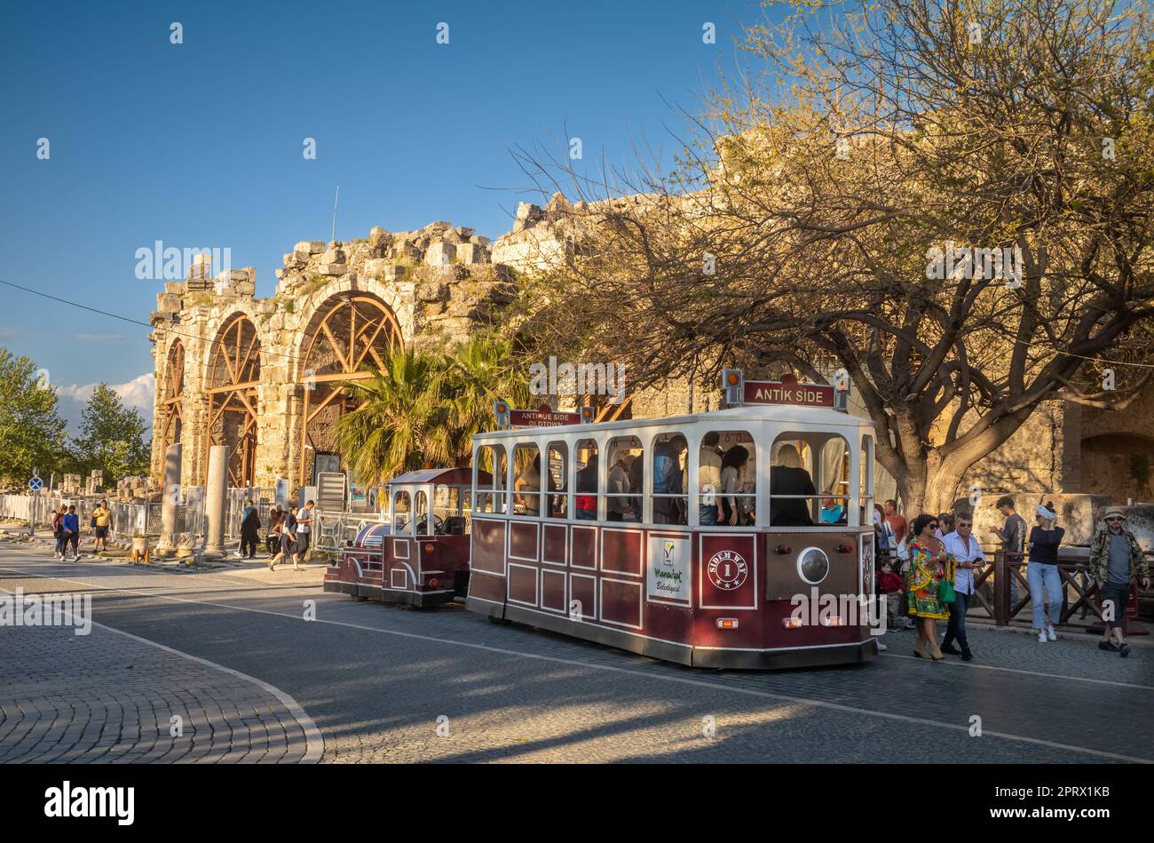 Touristen steigen vor den Ruinen des Amphitheaters in der antiken römischen Stadt Side in der Provinz Antalya, Türkei (Turkiye), in einen falschen Zug oder eine falsche Straßenbahn. Die Stockfoto