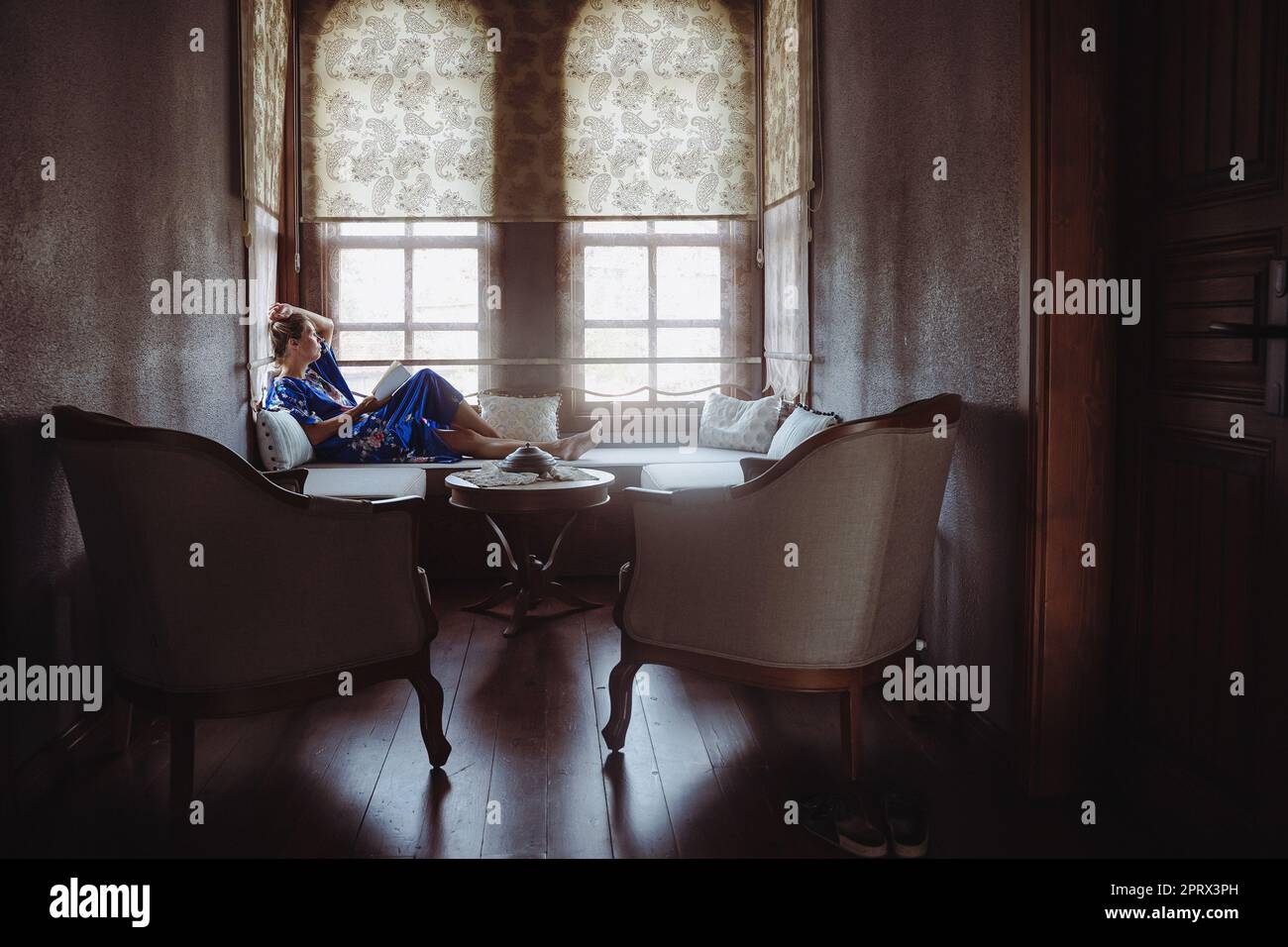 Frau in blauem Kleid, die sich auf dem Diwan mit mehreren Kissen entspannt und vor dem Fenster ein Buch in intimer Atmosphäre liest Stockfoto