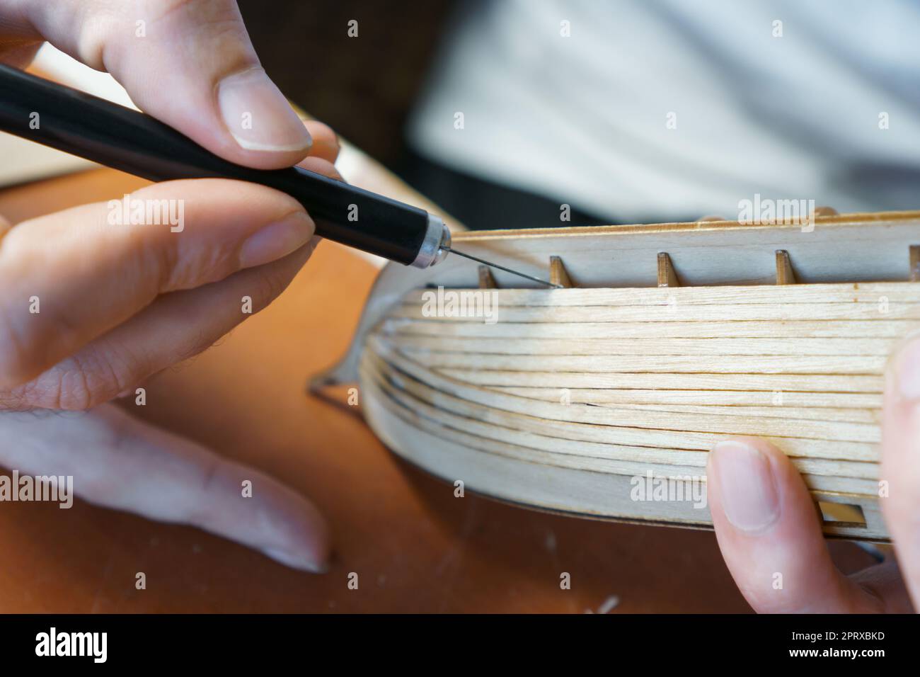 Hände des Mannes, der überschüssigen Klebstoff von Sperrholzdetails für Schiffsmodell mit Gebrauchsmesser entfernt. Prozess des Bauens Spielzeug Schiff, Hobby, Handwerk. Tabelle mit Stockfoto