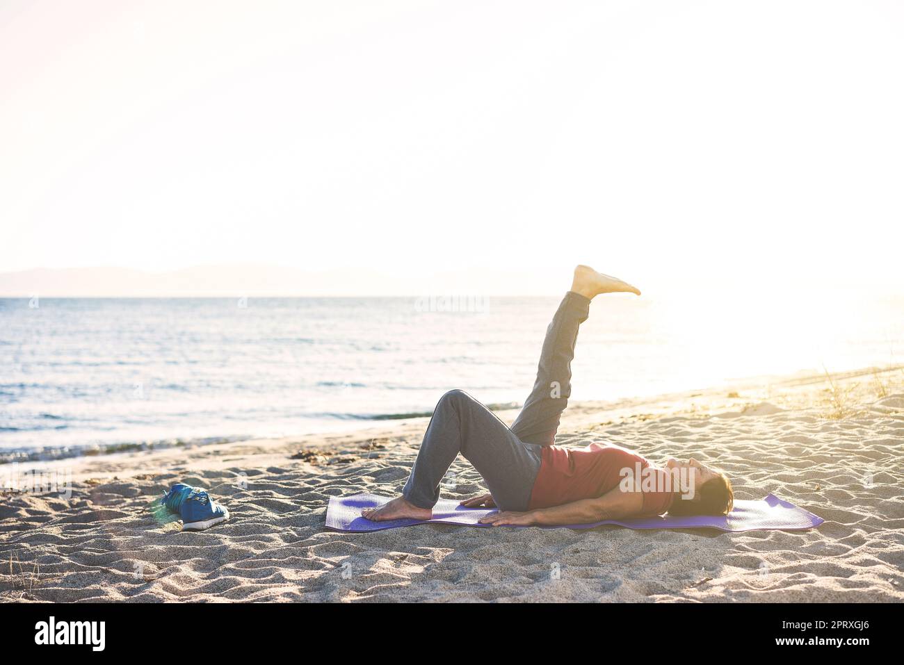 Seniorin am Strand liegt auf Yoga-Matte, trainiert. Beine heben. Stockfoto