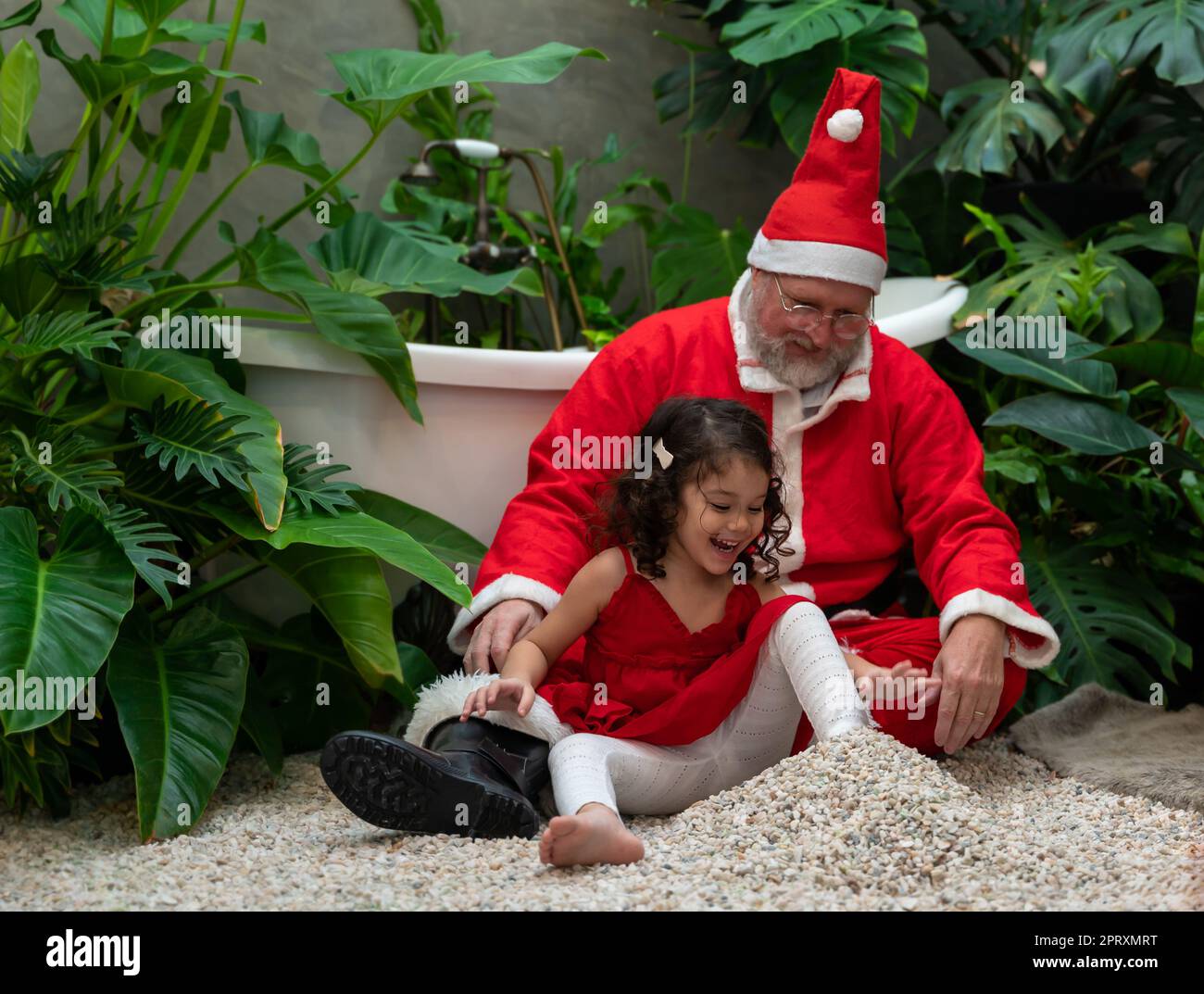 Fröhlicher Weihnachtsmann in rotem Kostüm und süßes kleines Mädchen in rotem Kleid, das auf Stein sitzt und zusammen spielt Stockfoto