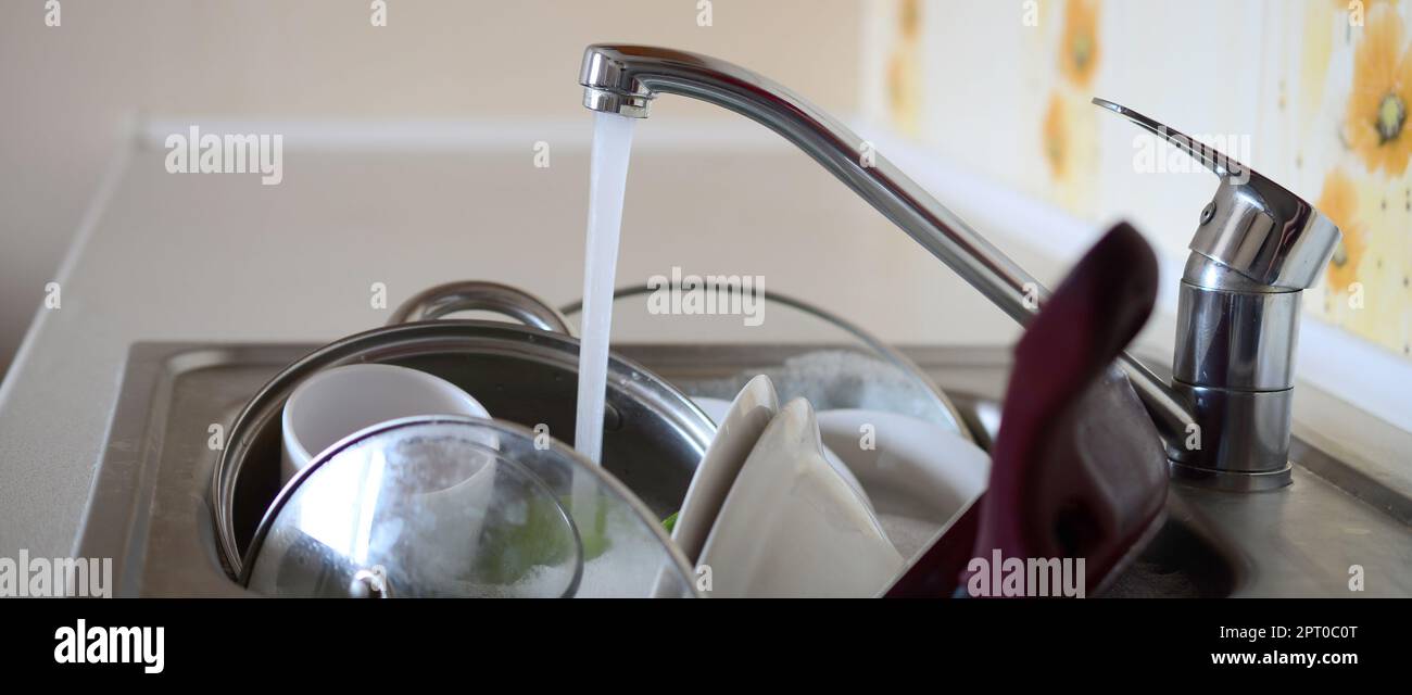 Schmutziges Geschirr und ungewaschene Küchengeräte liegen im Schaumwasser Unter einem Wasserhahn aus einer Küchenarmatur Stockfoto