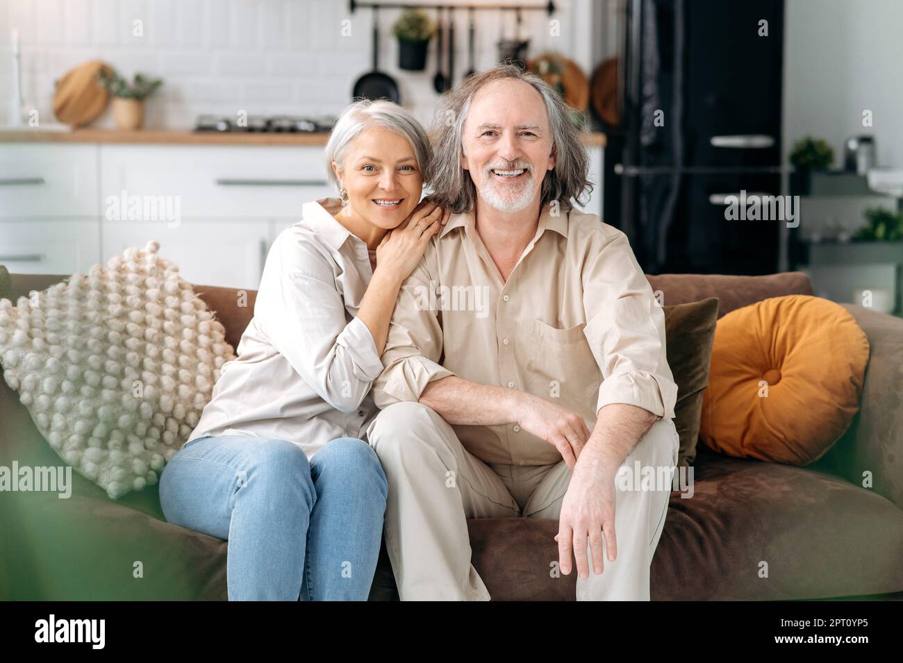 Porträt eines glücklichen Rentners. Reifes weißes Ehepaar, grauhaarige Ehepartner verbringen Zeit zusammen im Wohnzimmer zu Hause, Frau und Ehemann sitzen auf dem Sofa, umarmen sich, schauen in die Kamera, lächeln Stockfoto