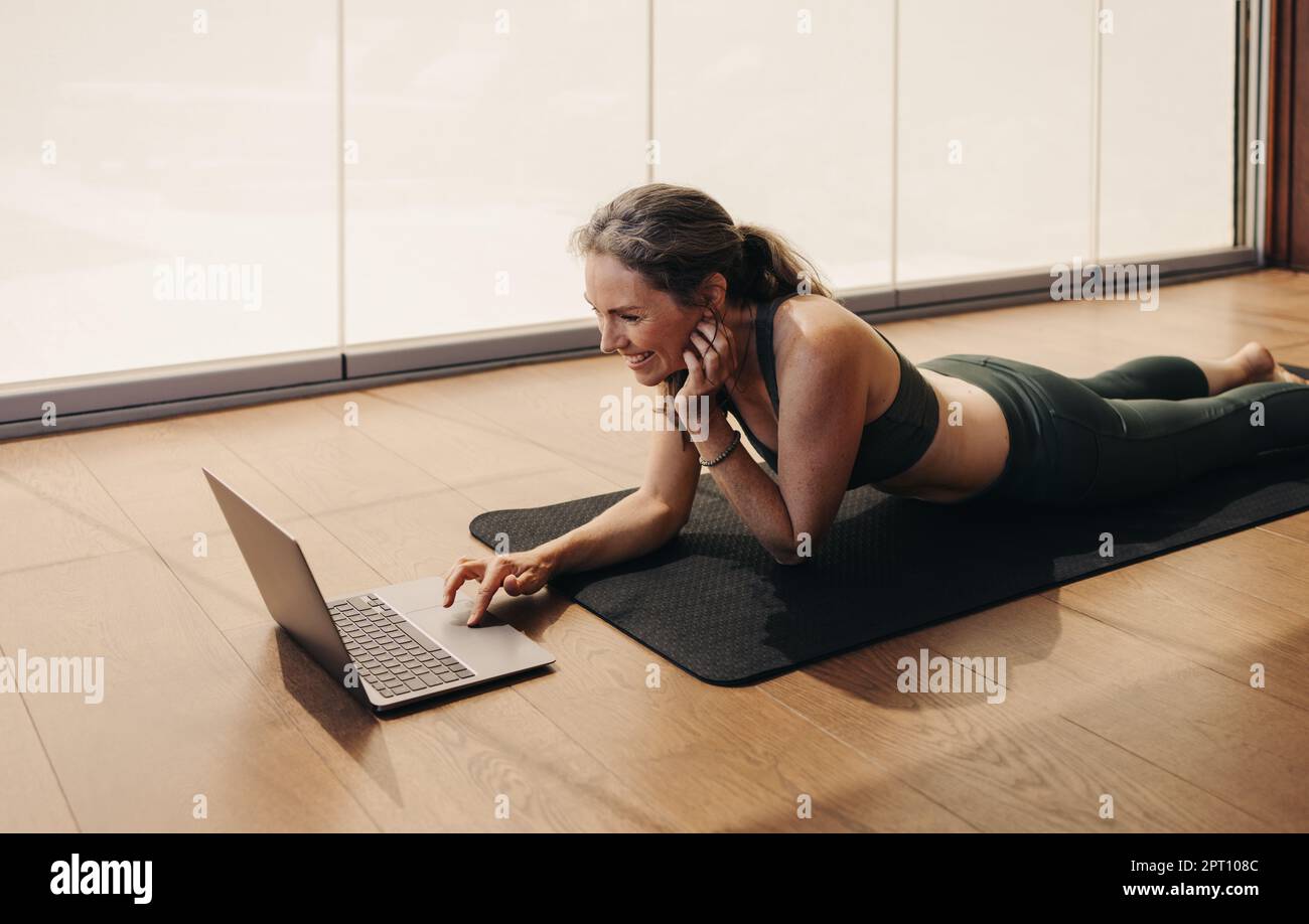 Ältere Frau lächelt, während sie an einem virtuellen Fitnesskurs auf einem Laptop teilnimmt. Glückliche Seniorin, die zu Hause ein Online-Yoga-Tutorial verfolgt. Fröhlich erwachsen Stockfoto