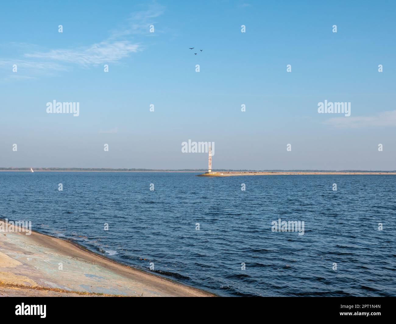 Ein alter BetonLeuchtturm am Rand des Kiew-Meeres spuckt mit Vögeln, ruhigem blauem Wasser mit einer weißen Yacht und blauem Himmel mit Federwolken Stockfoto