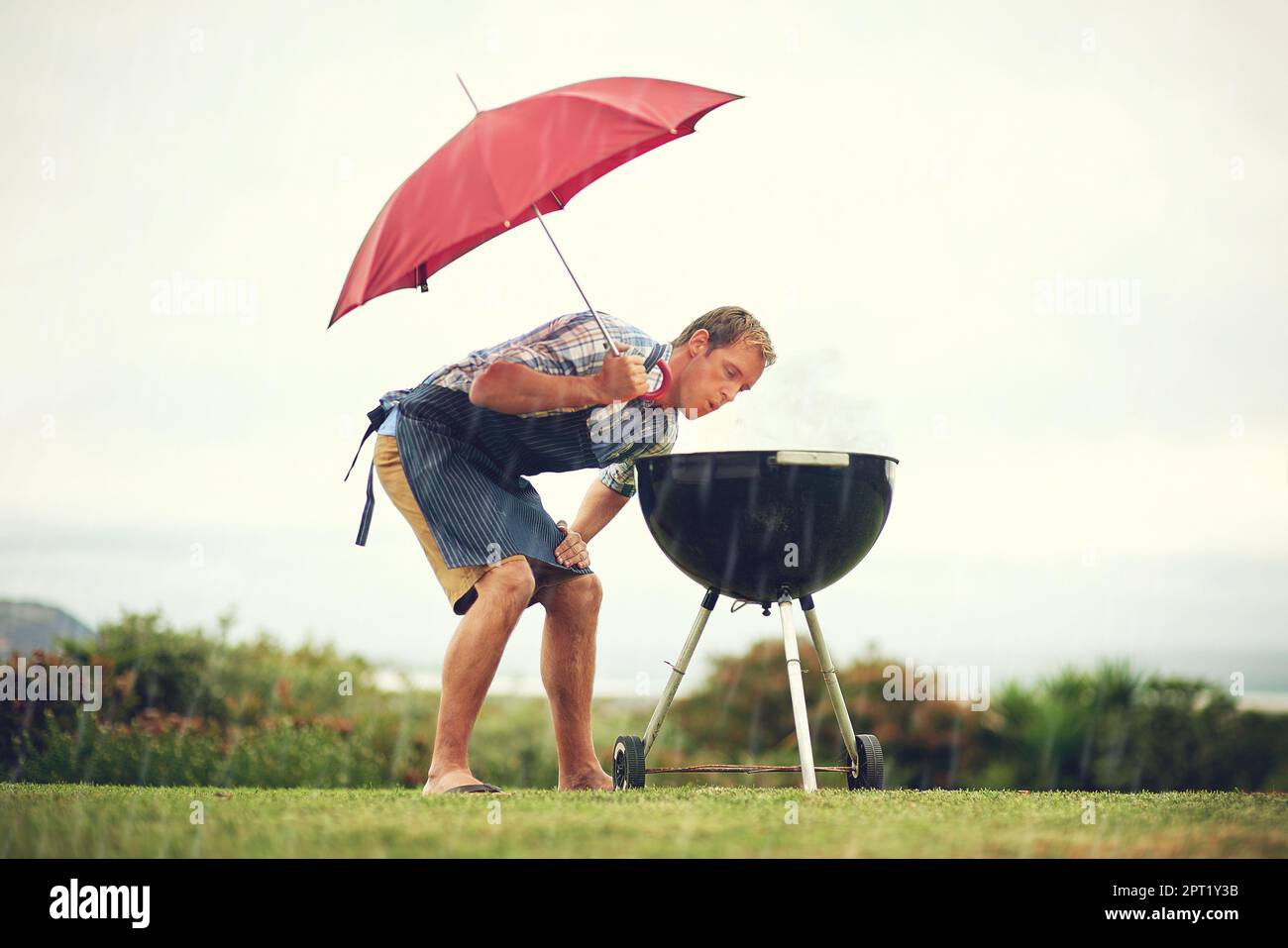 Wer sagt, Braaing ist immer leicht. Ein Mann, der draußen im Regen grillt, während er einen Schirm hält und das Feuer aufbläst Stockfoto