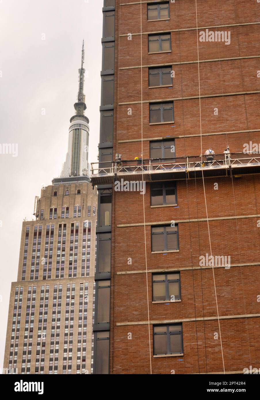 Die Arbeiter führen auf einer abgehängten Plattform Ziegelsteine mit dem Empire State Building im Hintergrund durch, 2023, New York City, USA Stockfoto