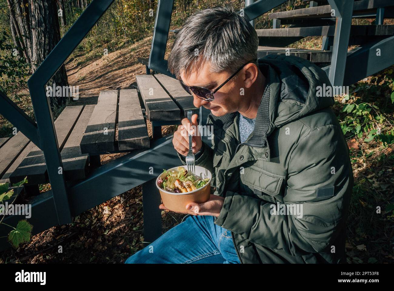 Ein Mann isst auf einem Salat aus einer Einwegpapierschüssel im Freien während eines Spaziergangs in der Natur im Herbst. Stockfoto