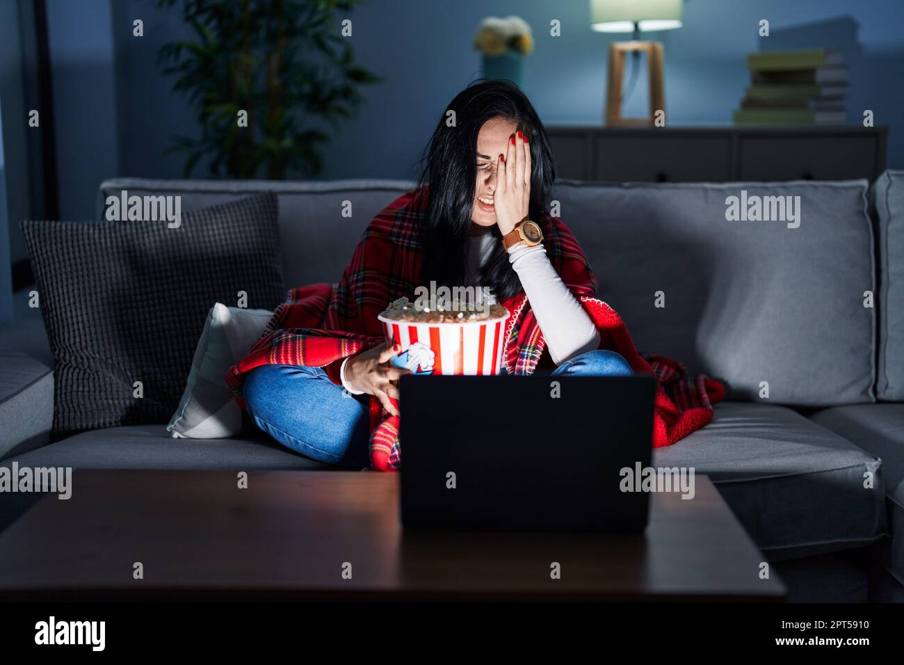 Hispanische Frau, die Popcorn isst und sich einen Film auf dem Sofa ansieht, und müde gähnt, indem sie das halbe Gesicht, die Augen und den Mund mit der Hand bedeckt. Das Gesicht tut weh. Stockfoto