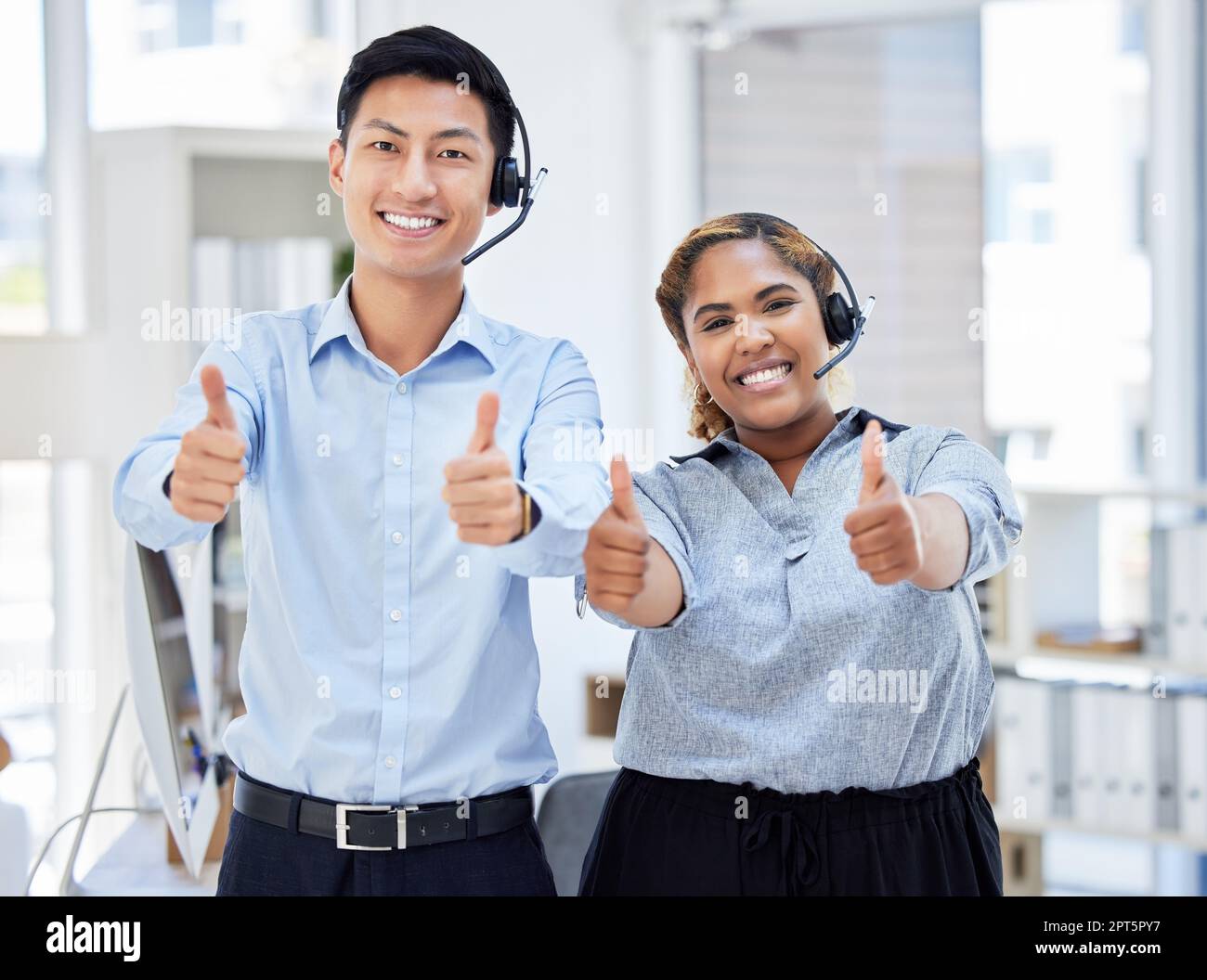 Zwei junge, glückliche, männliche und weibliche Callcenter-Agenten zeigen Daumen nach oben, während sie Anrufe in einem Büro beantworten. Zufriedener asiatischer Mann und gemischter Stockfoto