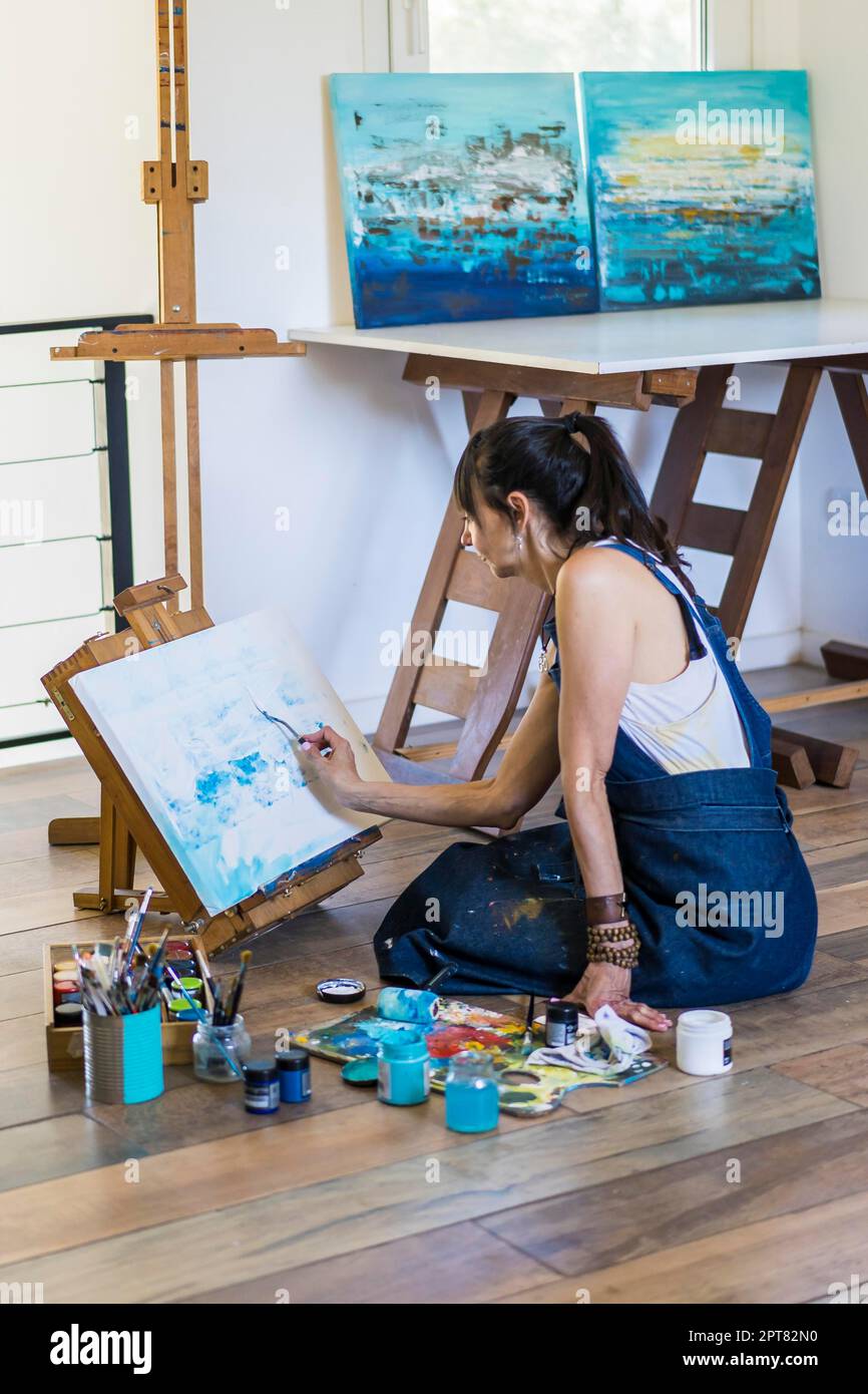 Künstlerin malt auf einer Leinwand ein blaues abstraktes Gemälde. Kreative Frau, die in ihrem Kunstatelier auf dem Boden arbeitet Stockfoto