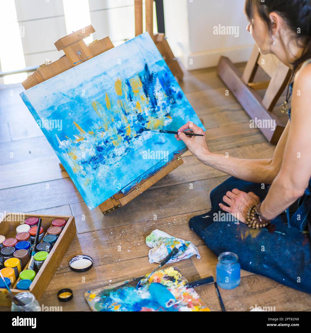 Künstlerin malt auf einer Leinwand ein blaues abstraktes Gemälde. Kreative Frau, die in ihrem Kunstatelier auf dem Boden arbeitet Stockfoto