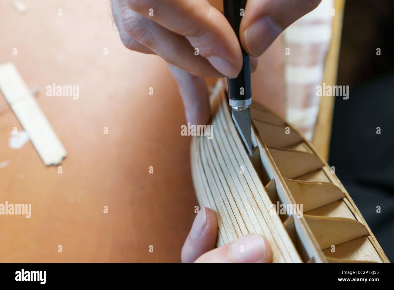 Hände des Mannes, der überschüssigen Klebstoff von Sperrholzdetails für Schiffsmodell mit Gebrauchsmesser entfernt. Prozess des Bauens Spielzeug Schiff, Hobby, Handwerk. Tabelle mit Stockfoto