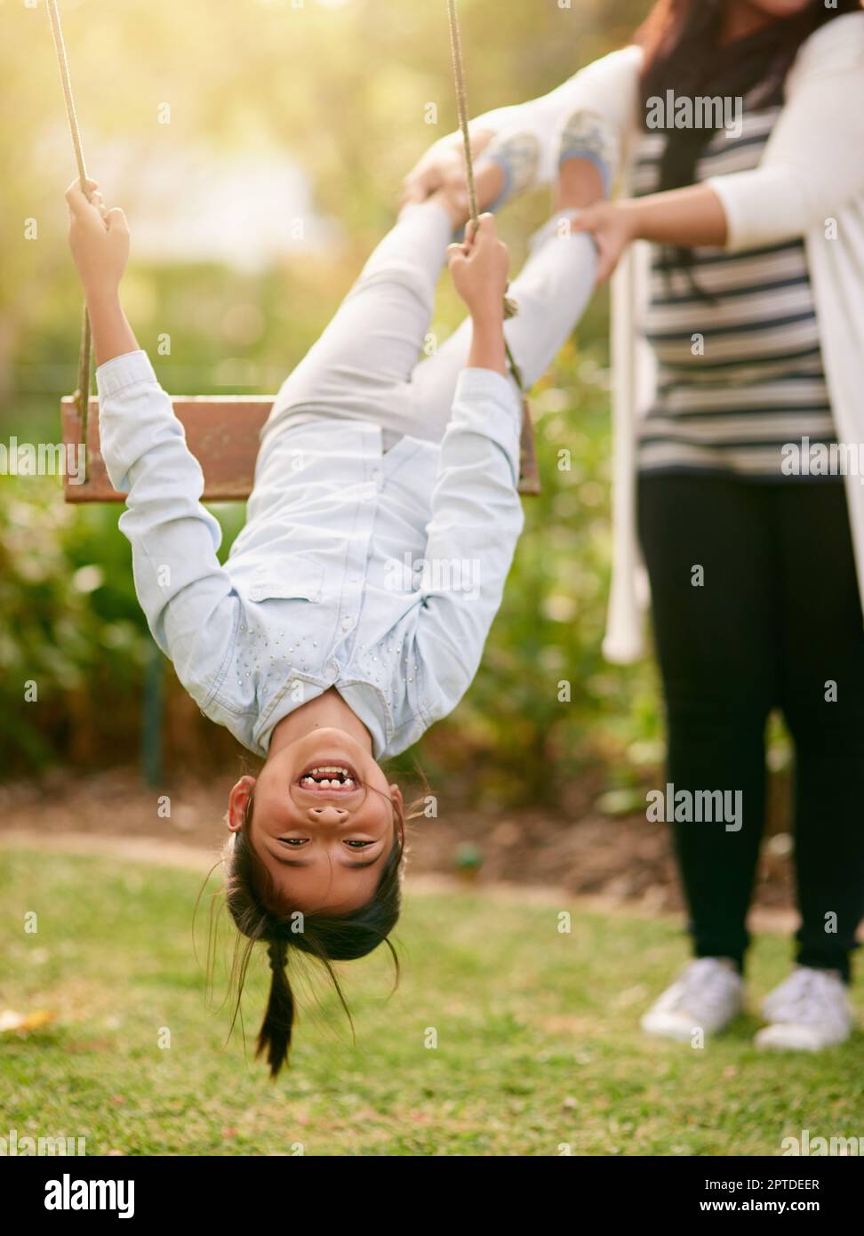 Nach oben, nach unten. Porträt eines kleinen Mädchens, das draußen mit seiner Mutter im Hintergrund auf einer Schaukel spielt Stockfoto
