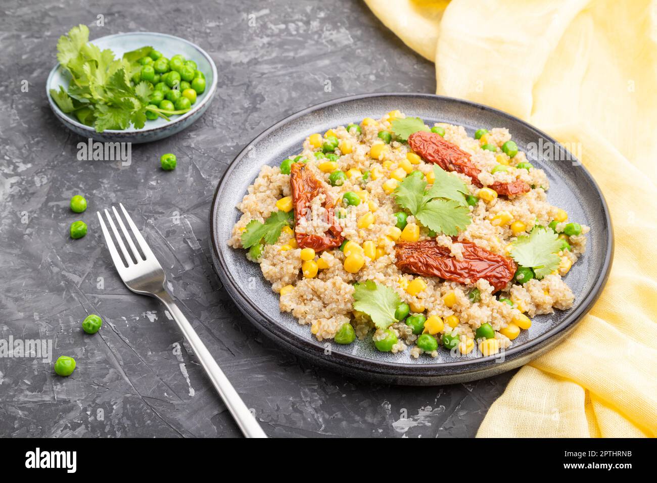 Quinoa-Haferbrei mit grüner Erbse, Mais und getrockneten Tomaten auf Keramikplatte auf grauem Betongrund und gelbem Textil. Seitenansicht, Nahaufnahme. Stockfoto