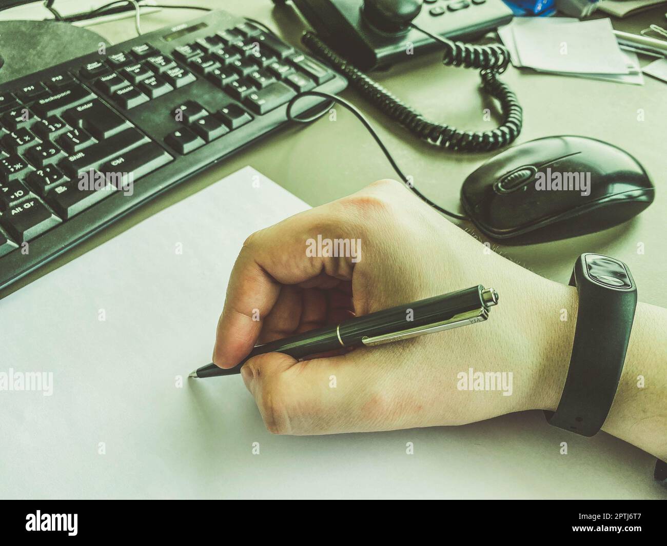 In der Hand des Büromitarbeiters befindet sich ein schwarzer Metallstift mit einer Stange. Handschriftliche Notizen auf einem weißen Stück Papier machen. Ein Mann, der neben einem Computer schreibt Stockfoto