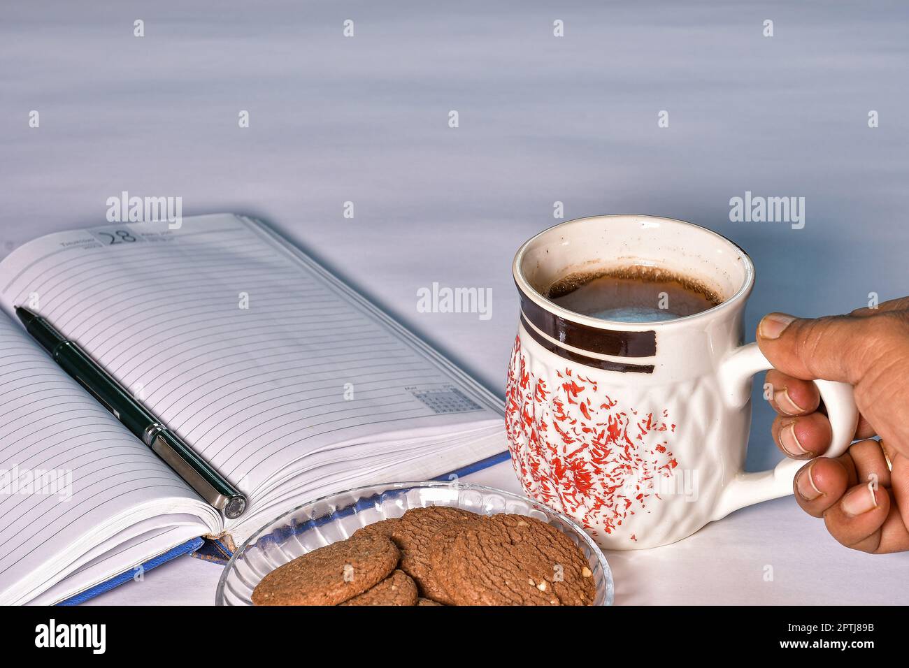 Eine Hand nimmt eine Tasse schwarzen Tee vom Arbeitstisch, um sie zusammen mit einem Teller Kekse und einem Tagebuch mit Stift zu genießen - ideal für Werbung Stockfoto