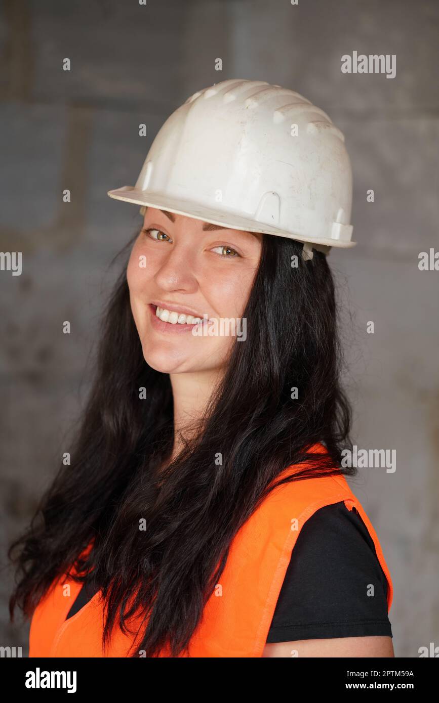 Junge Frau in weißem Schutzhelm und orangefarbener Warnjacke, langes dunkles Haar, schaut in die Kamera und lächelt. Nahaufnahme von Detail zu Gesicht, verschwommene Kontras Stockfoto