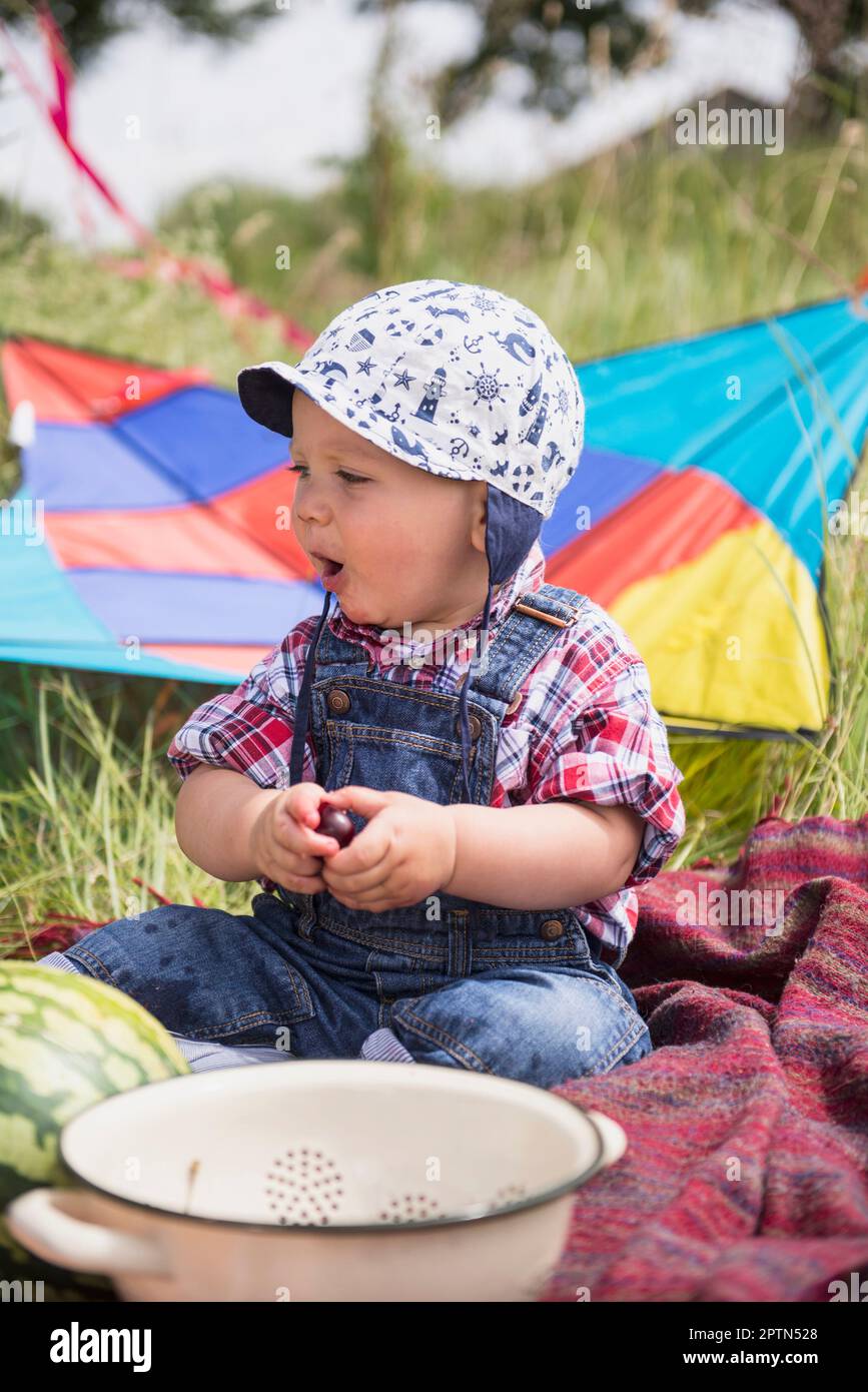 Kleiner Junge mit Kirsche, der auf einer Picknickdecke auf dem Land sitzt, Bayern, Deutschland Stockfoto