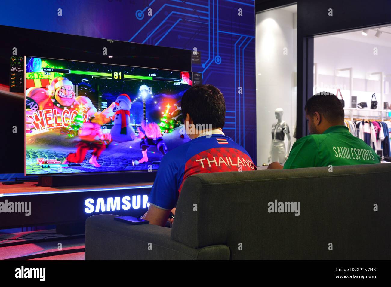 Zwei Männer sitzen und spielen ein Videospiel auf einem großen Samsung-Fernseher in der Central Festival Mall, Pattaya, Thailand. Stockfoto