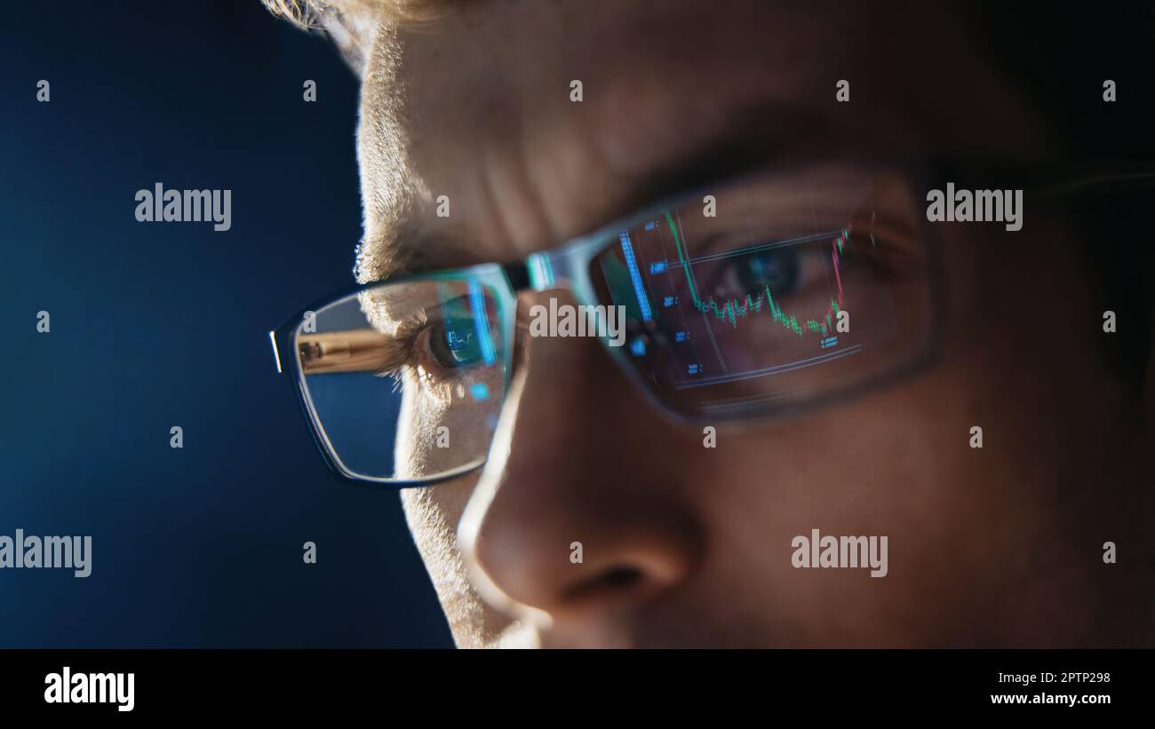 Nahaufnahme eines fokussierten Händlers trägt eine Brille, sieht sich den  pc-Bildschirm an, wobei die Aktienkurven reflektiert werden, und arbeitet  nachts online. Ein Mann, der den Krypto-Markt analysiert. Selektiver Fokus  auf das Auge.