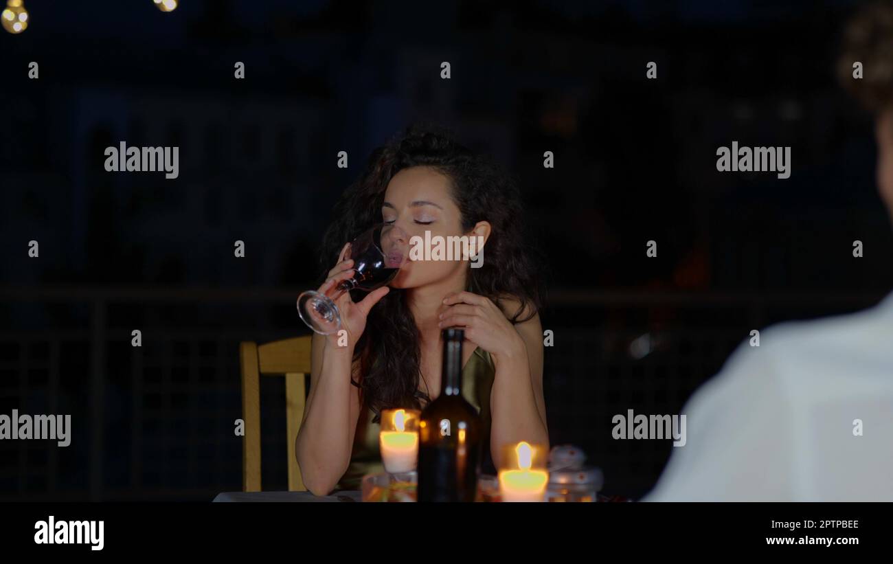 Porträt einer kaukasischen Dame in Kleidern, die abends draußen mit einem Glas Wein beim Date sitzt und spricht. Zeitlupe. Romantisches Beziehungskonzept. Ein Paar Stockfoto