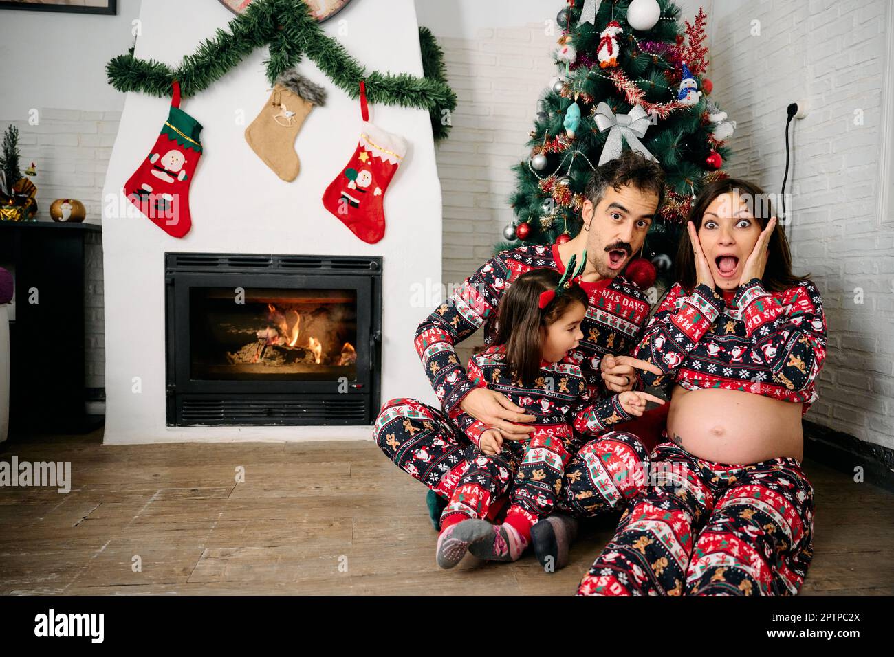 Eine Familie, mit Vater, Mutter und Tochter, die dazu passende Weihnachtsschlafanzüge tragen, Familienfotos vor dem Kamin machen und einen Weihnachts-Tr Stockfoto