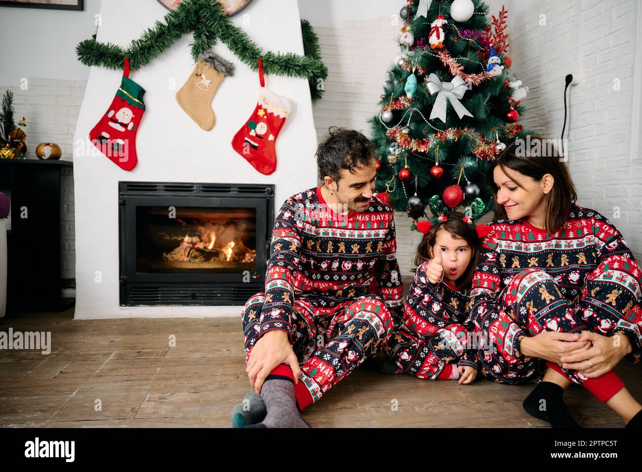 Eine Familie, mit Vater, Mutter und Tochter, die dazu passende Weihnachtsschlafanzüge tragen, Familienfotos vor dem Kamin machen und einen Weihnachts-Tr Stockfoto