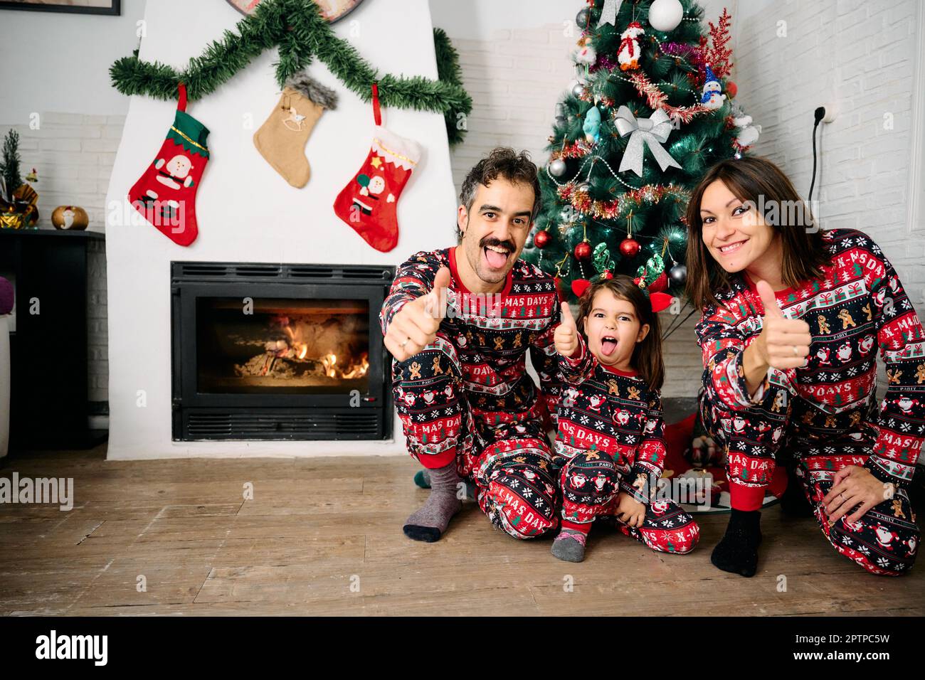 Eine Familie, mit Vater, Mutter und Tochter, die dazu passende Weihnachtsschlafanzüge tragen, Familienfotos vor dem Kamin machen und einen Weihnachts-Tr Stockfoto